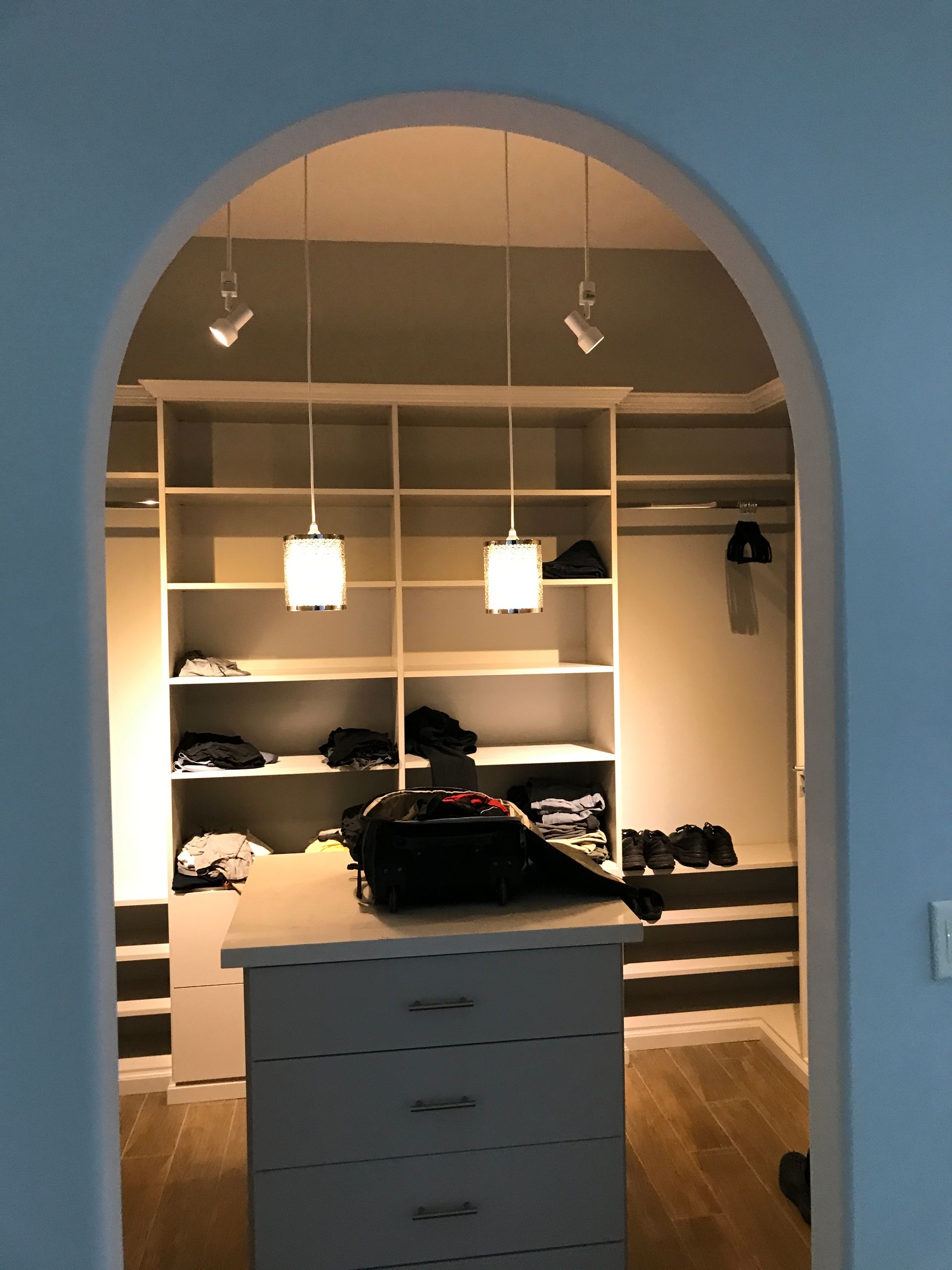 Walk-in closet with built-in shelves and drawers, lit by hanging lights. Features a central island, items on shelves.
