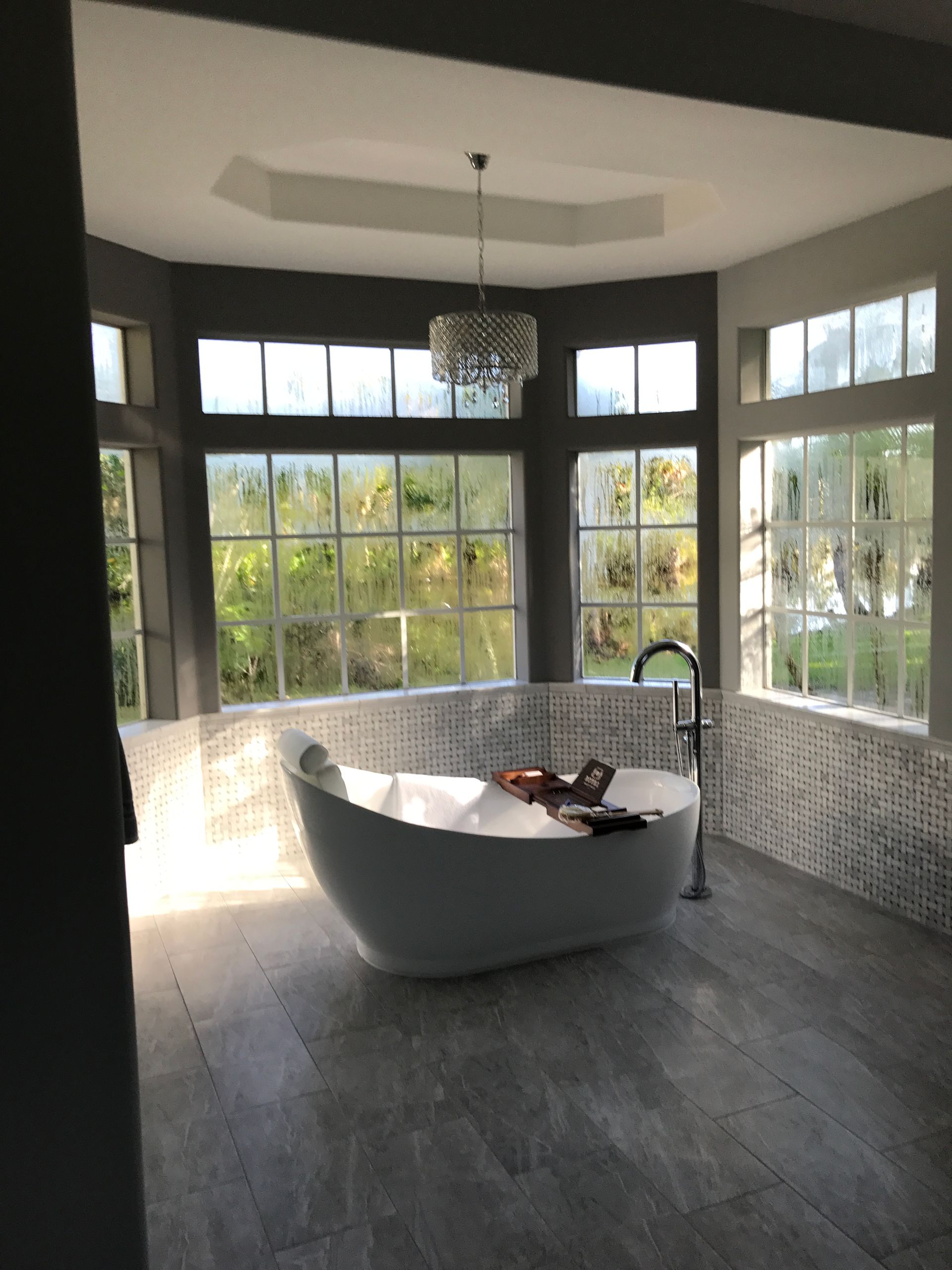 A modern bathroom with a freestanding tub by a window, gray floors and walls, and a chandelier.