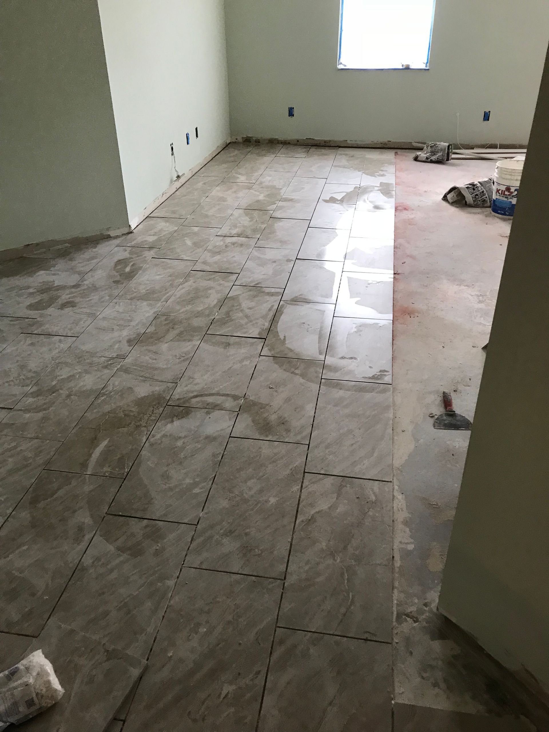 Room with a partially tiled floor, construction in progress. Unfinished walls, window, tools, and debris are visible.