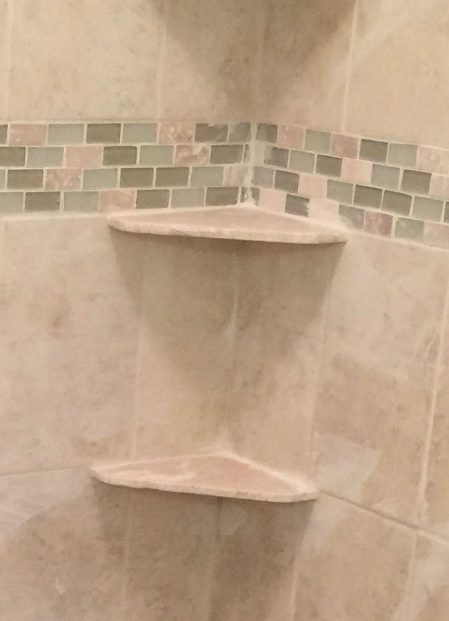 Shower corner with two built-in, light-colored shelves and decorative tile band.