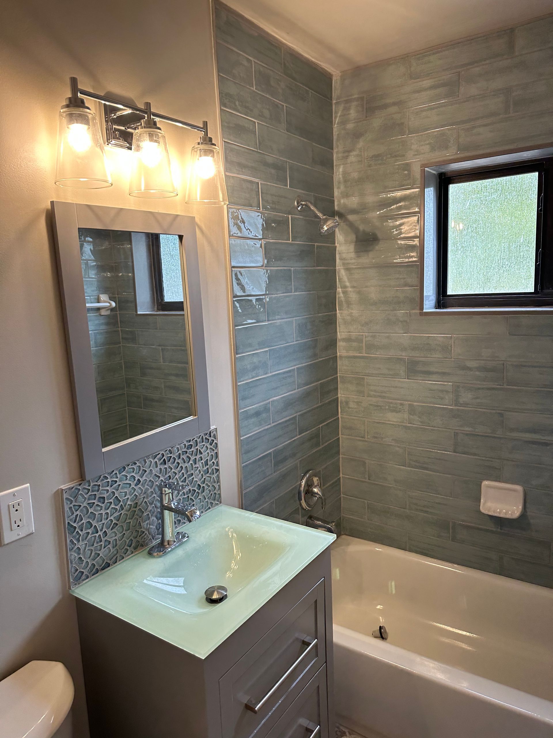 Bathroom with gray vanity, blue-gray tiled shower, white tub, and window.