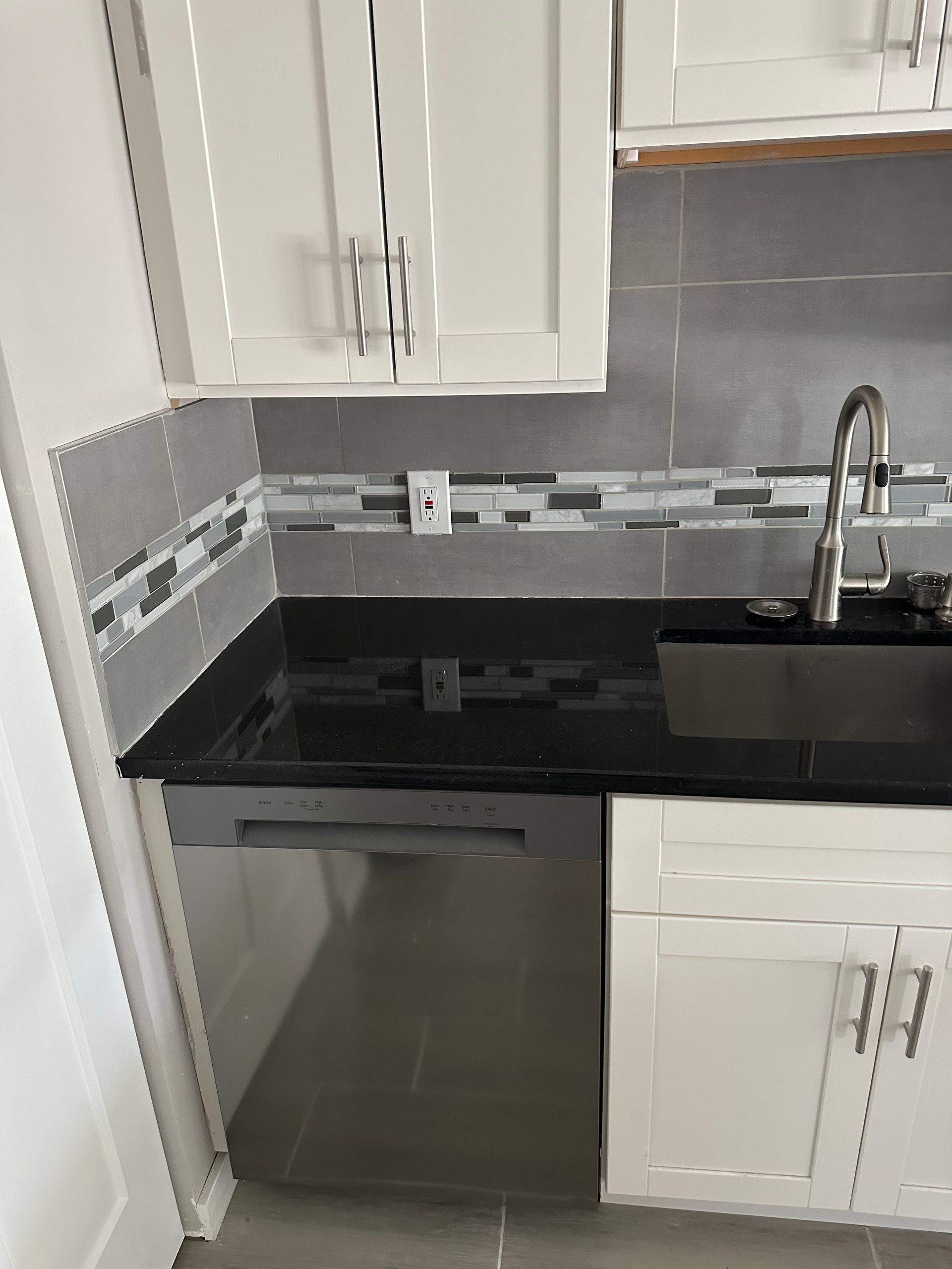 Kitchen with white cabinets, black countertop, silver dishwasher, and tile backsplash.