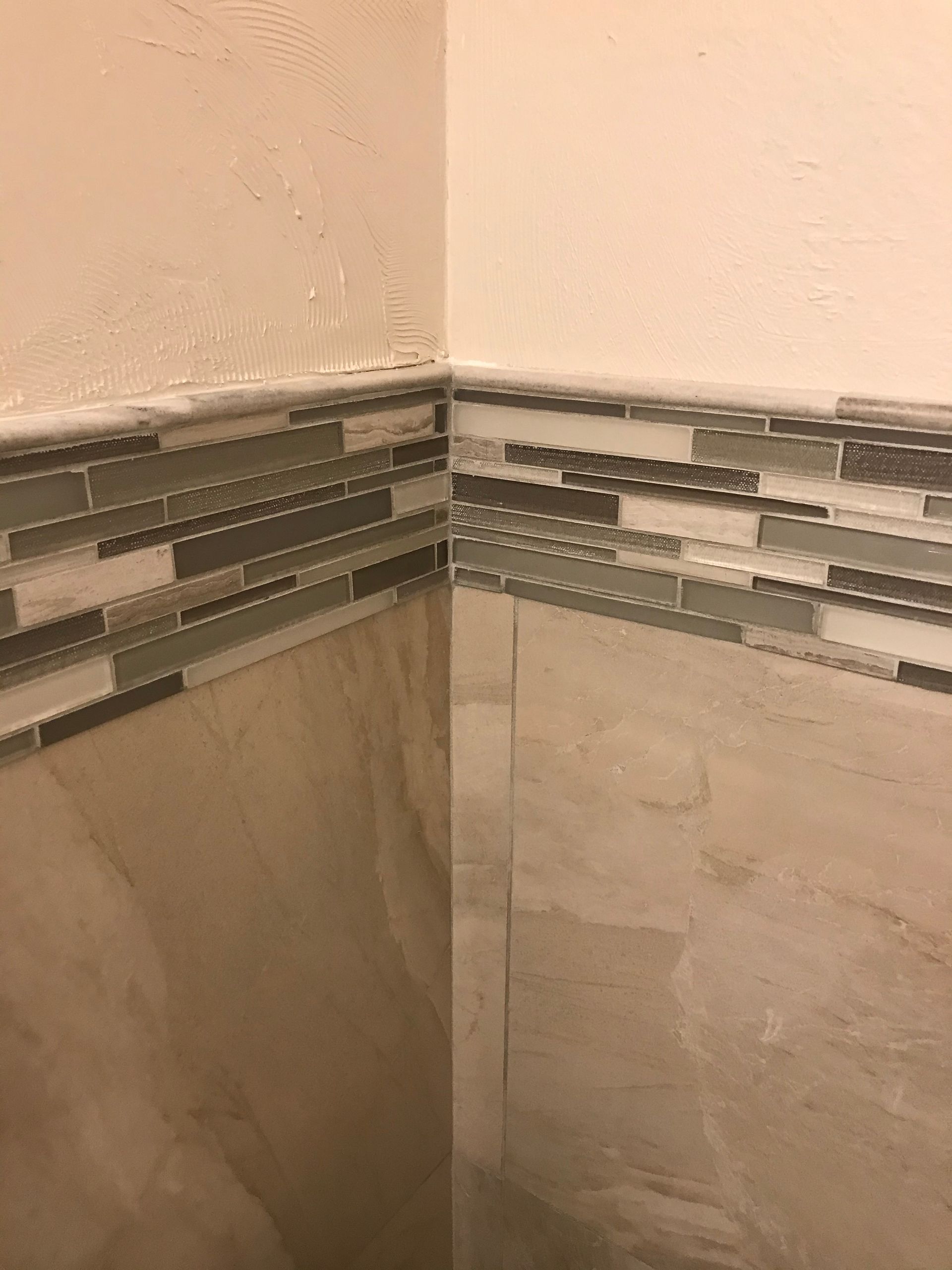 Corner of bathroom with decorative tile border, beige wall and large tile, white wall.