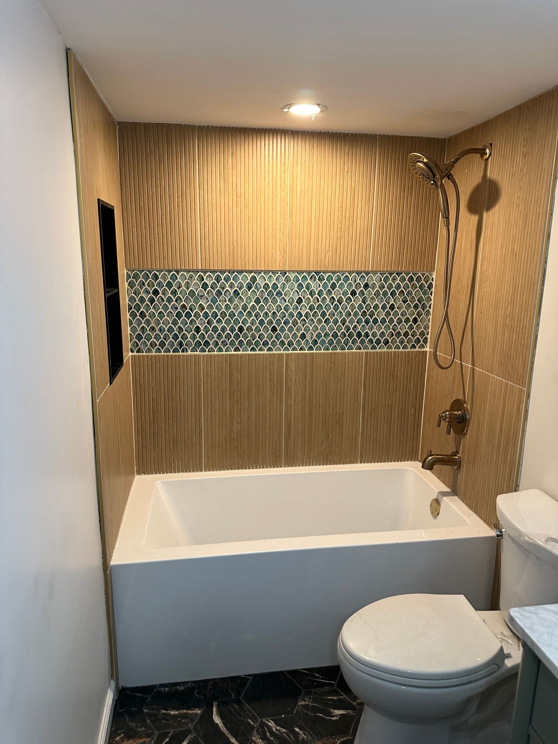 Bathroom with white tub and toilet. Beige ribbed tile with teal accent tile. Antique brass shower fixtures.