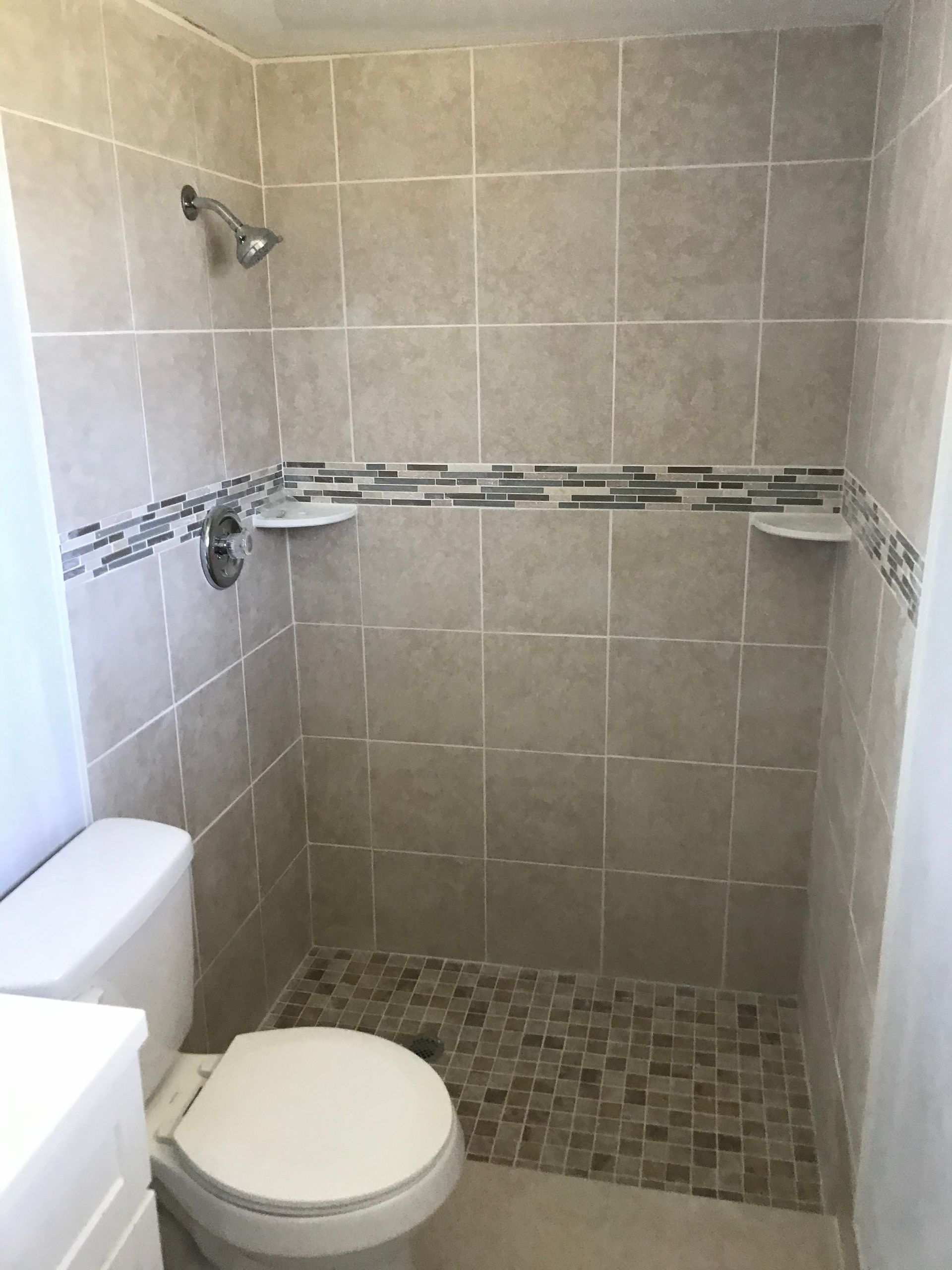 Beige-tiled shower with mosaic accent, two shelves, showerhead, and toilet.