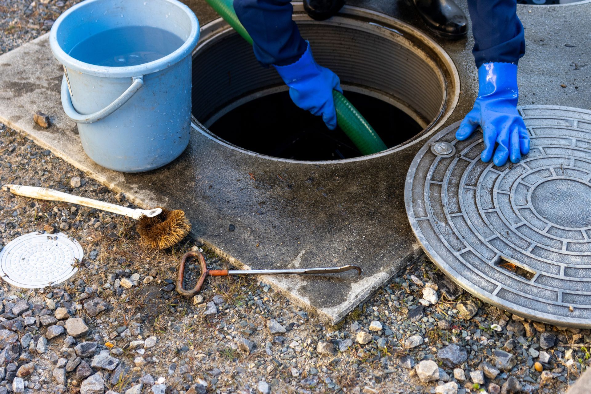 A person in blue protective gloves uses a green hose to clean an open manhole, with a bucket and tools nearby.