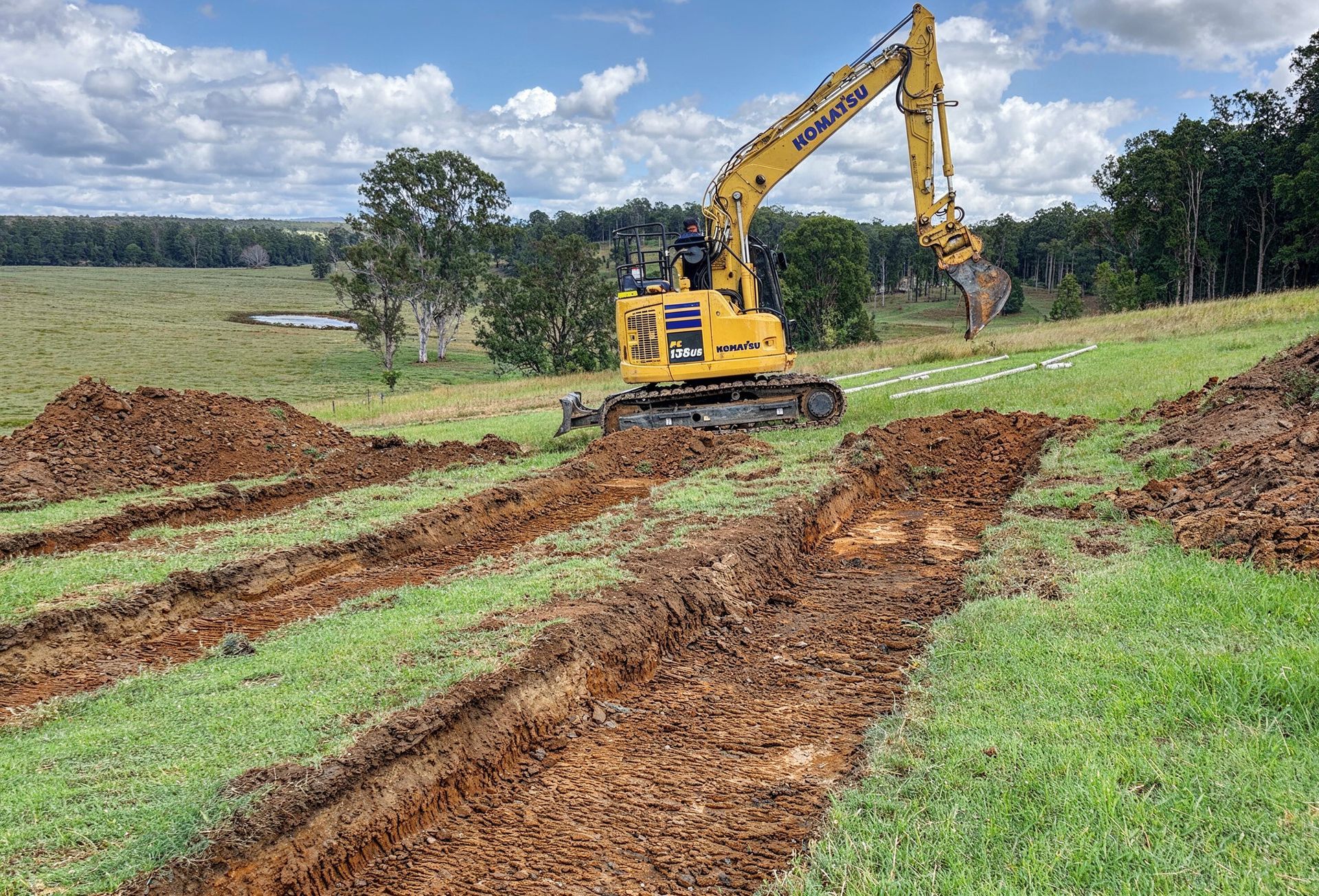 A yellow excavator sits on a grassy hillside, having dug several long trenches in the dirt.