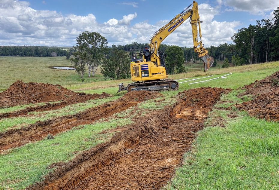 A yellow excavator sits on a grassy hillside, having dug several long trenches in the dirt.