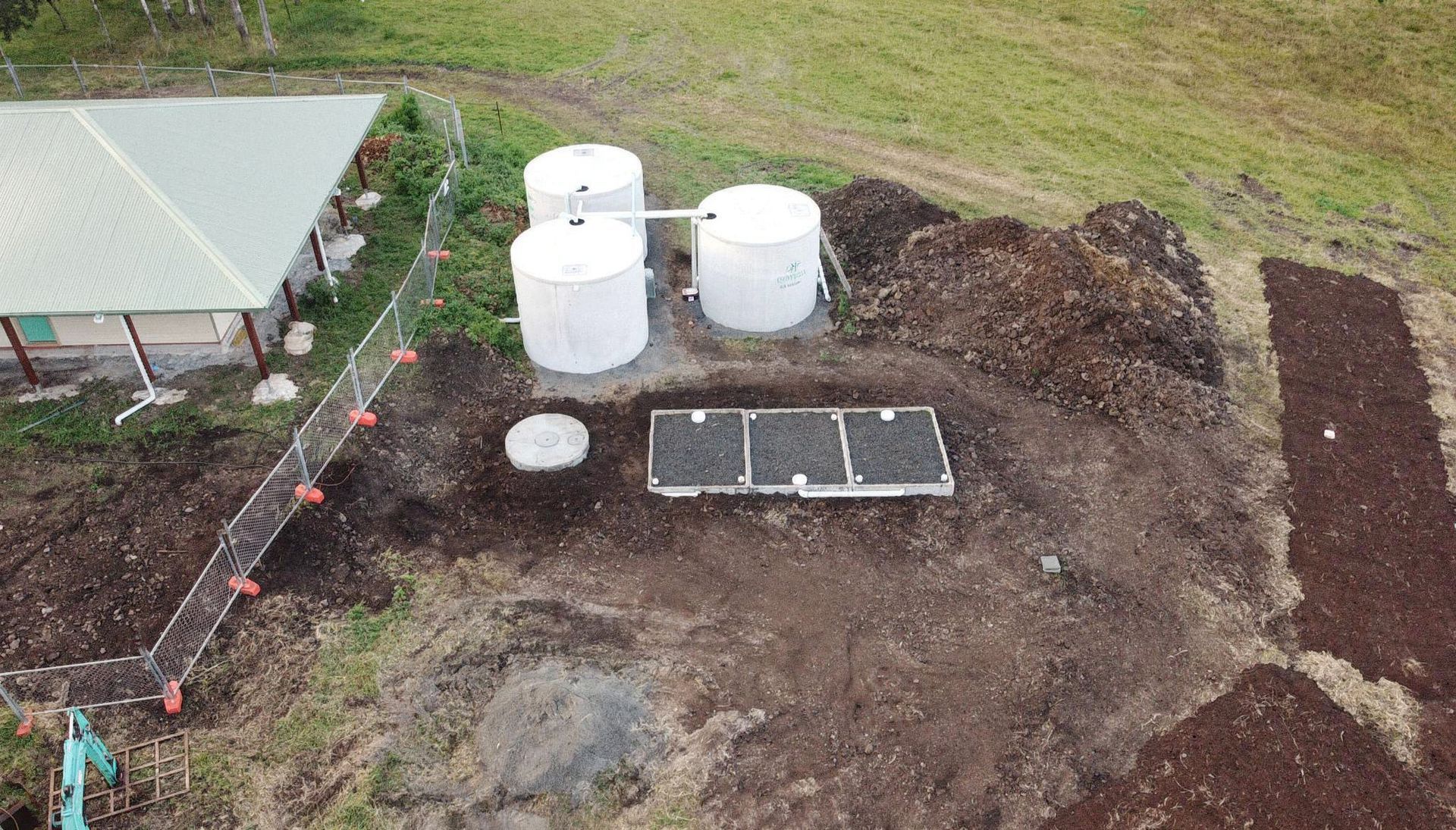 An aerial view shows three white water tanks, a concrete septic system, and a small building set in a grassy landscape.