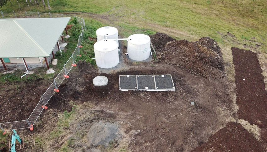 An aerial view shows three white water tanks, a septic system access cover, and a gravel filtration bed in a yard.