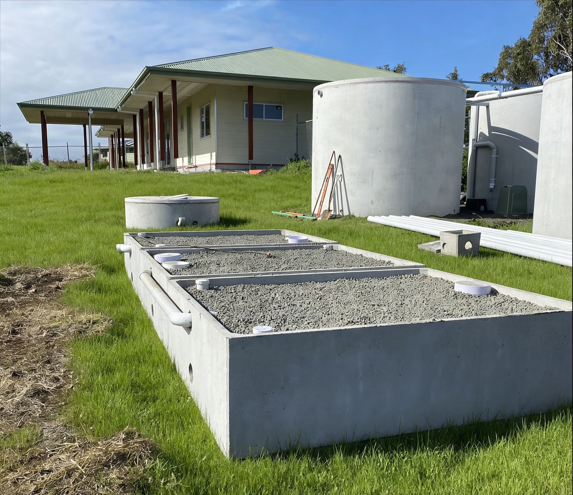 A rectangular concrete septic gravel filter next to large water tanks on a grassy lawn near a house.