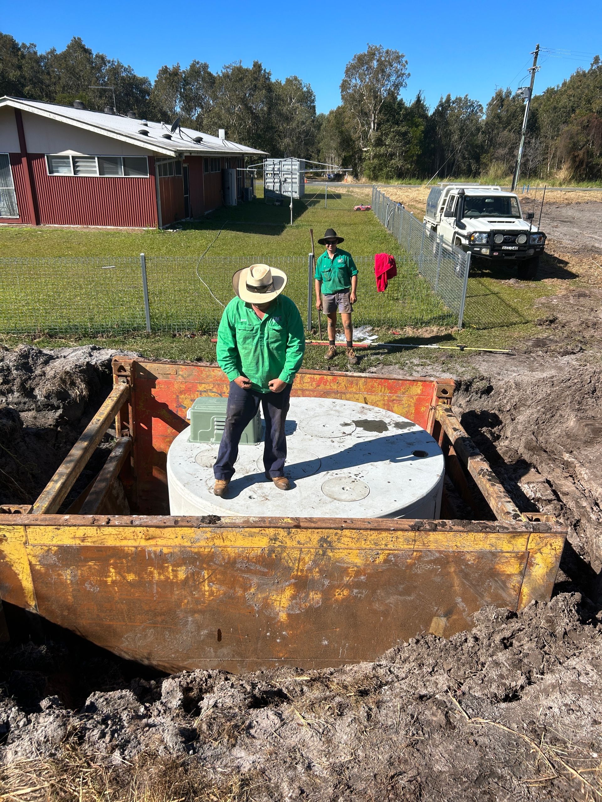 Two people in hats and work shirts stand by a large, circular concrete structure inside an open trench on a sunny day.