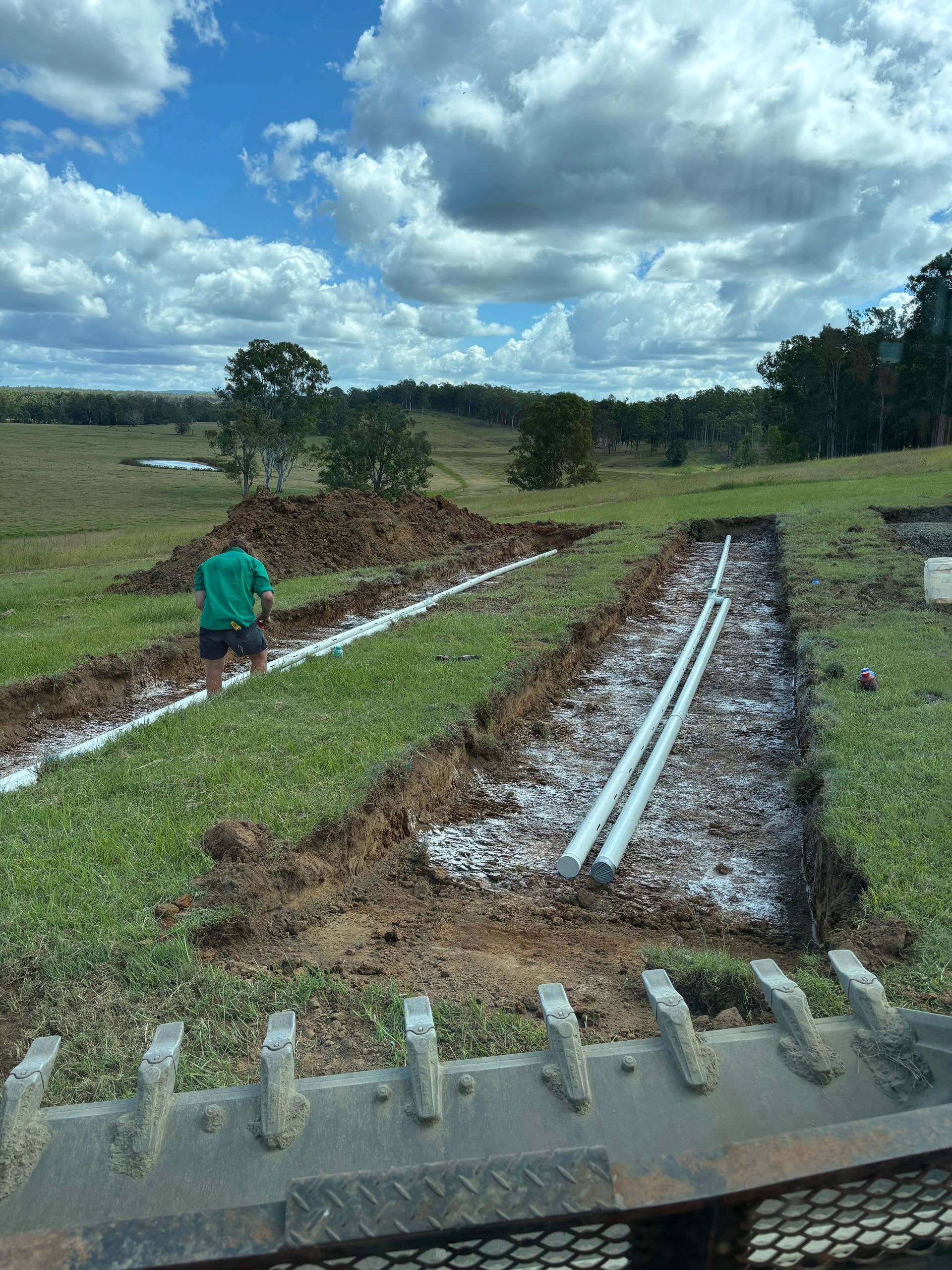 A person stands in a grassy field near two trenches containing drainage pipes and gravel, viewed from behind an excavator.