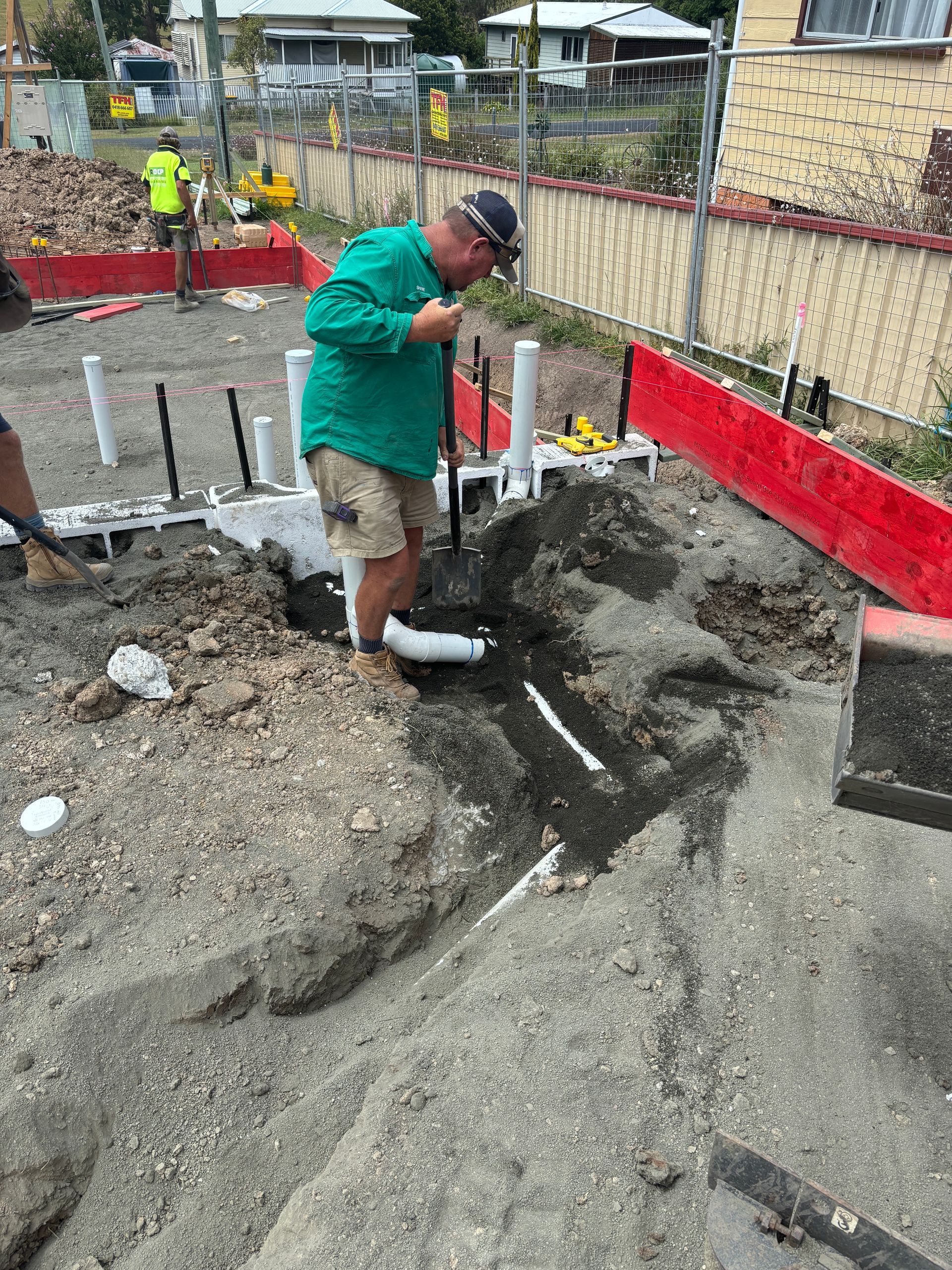 A worker in a green shirt digs into the dirt at a construction site with white pipes protruding from the ground.