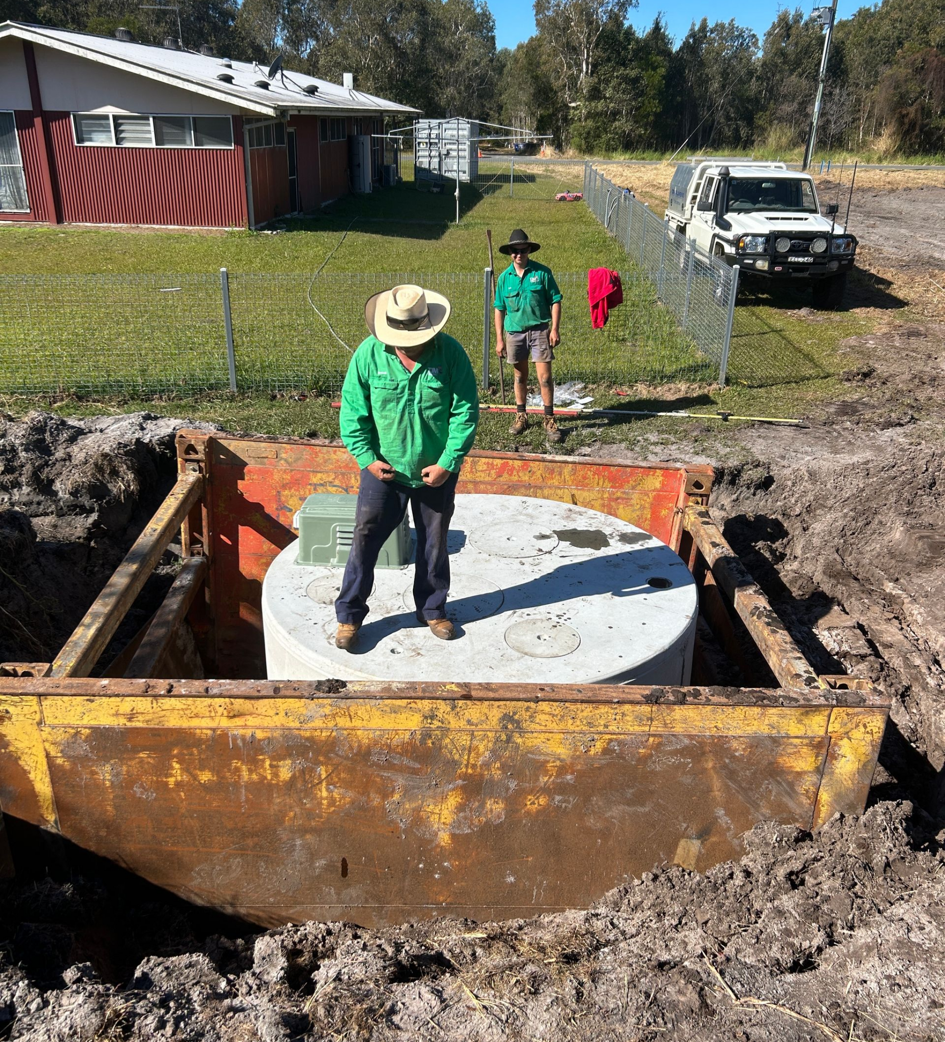 A person in a green shirt and hat stands on a round concrete structure inside a deep trench, while another person watches.