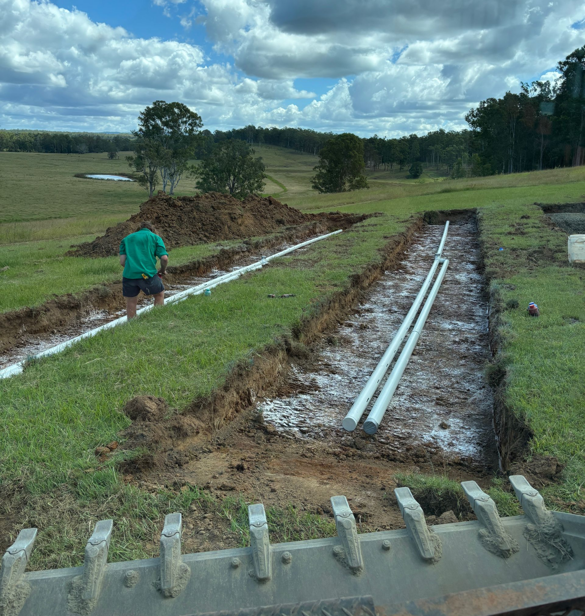 A person stands in a grassy field near two trenches containing white drainage pipes and gravel, viewed from an excavator.
