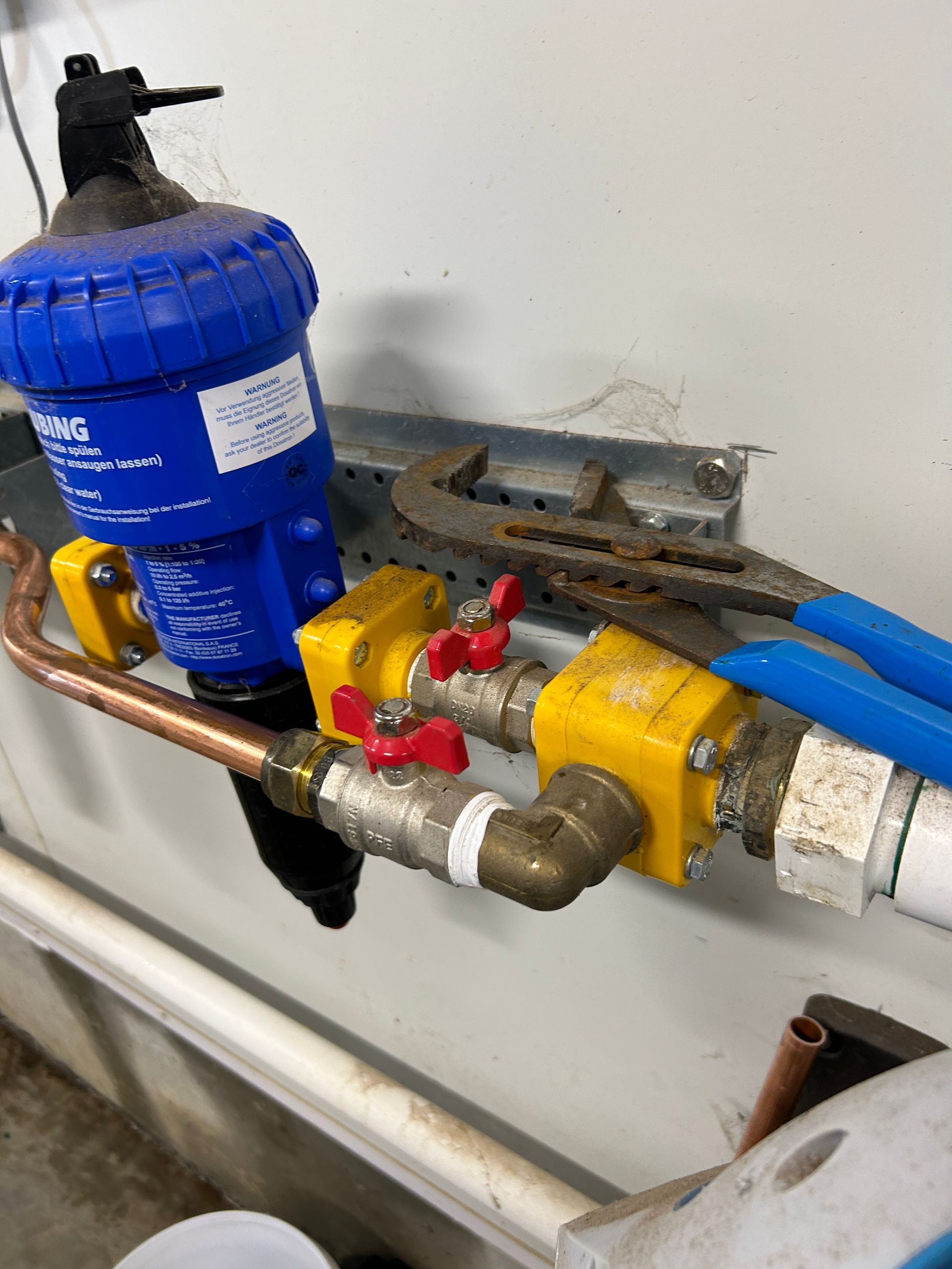 A water filtration system with yellow valves and copper piping mounted on a wall, with a pair of pliers resting on top.