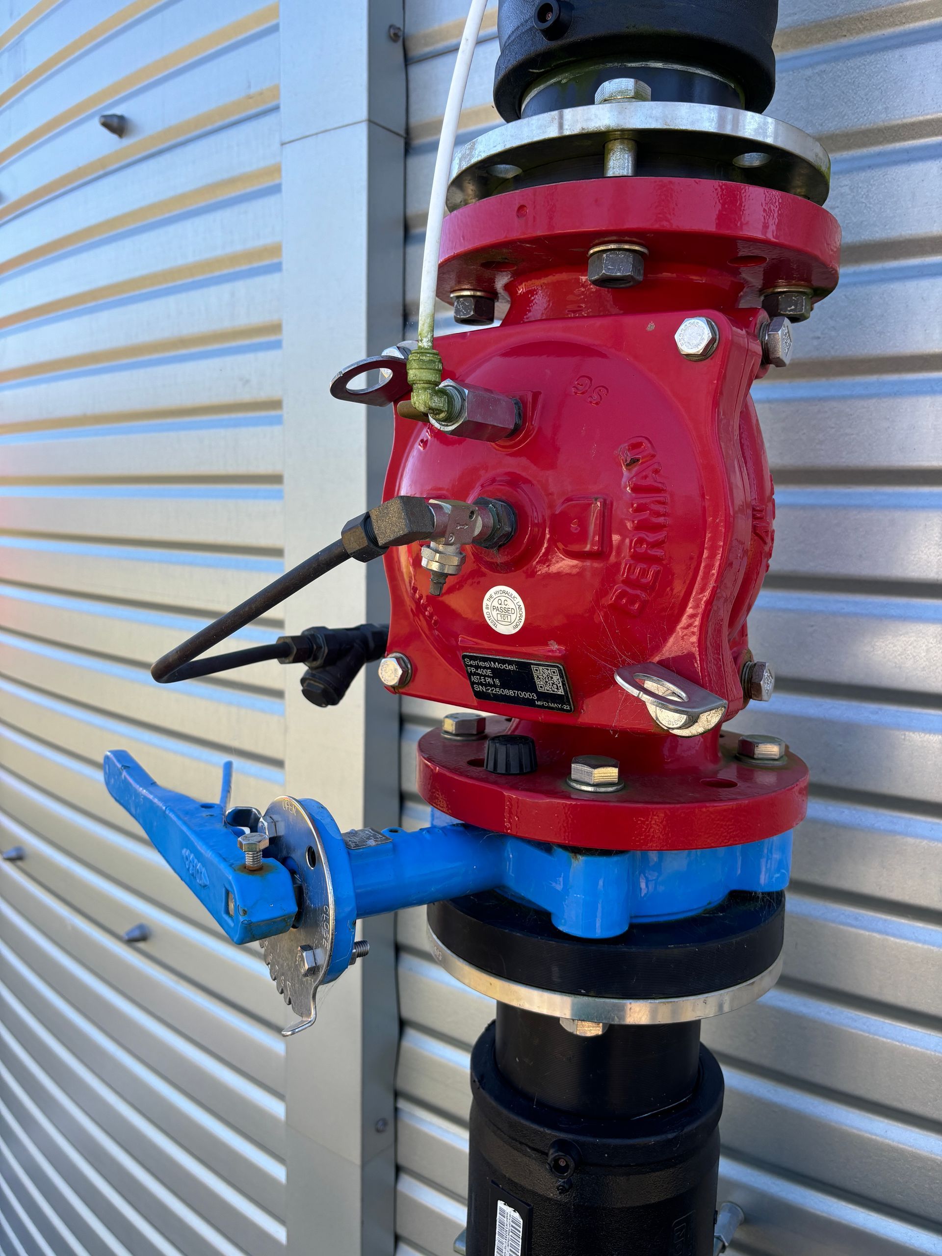 A red valve assembly with a blue handle attached to a vertical pipe against a corrugated metal wall.