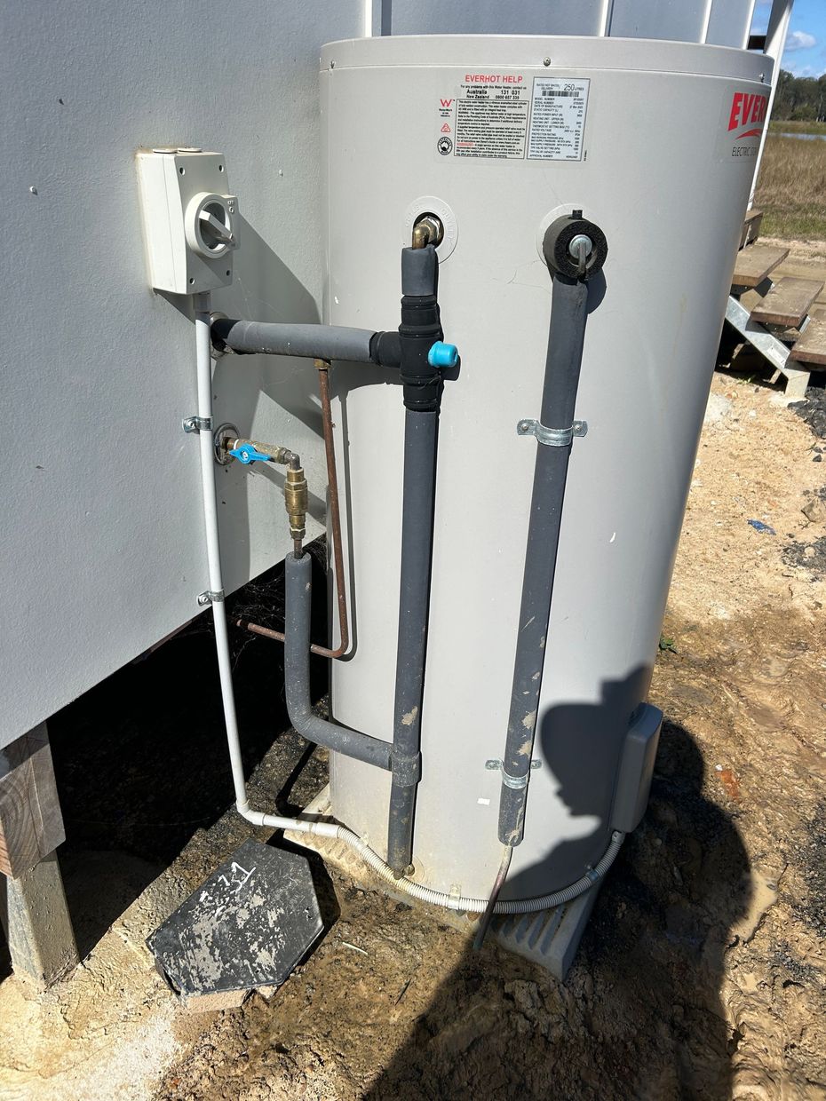 A gray electric hot water tank stands outdoors next to a building wall with connected water pipes and an electrical box.