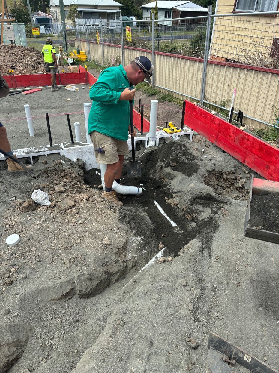 A construction worker in a green shirt digs a trench in the dirt of a residential building site.