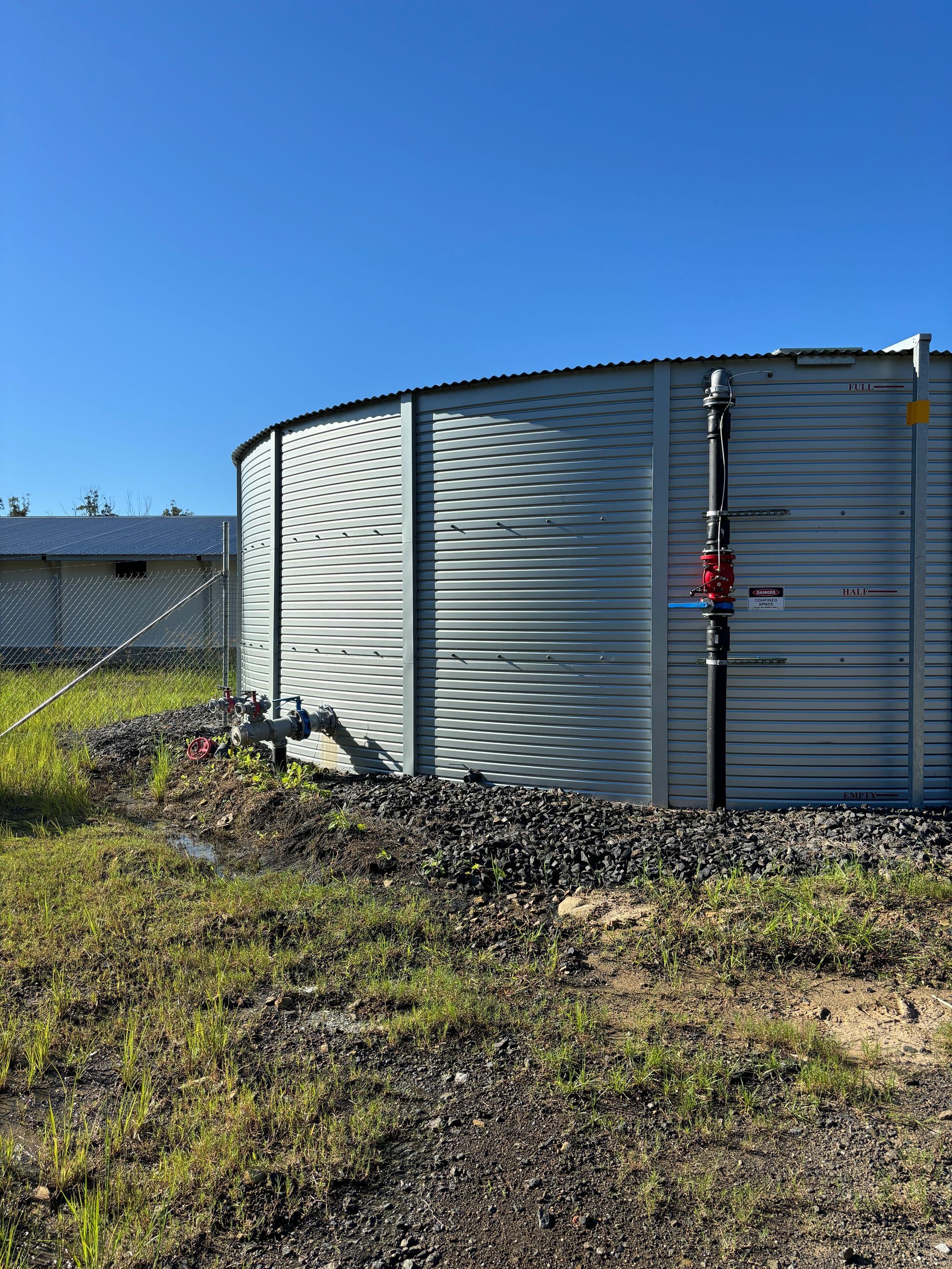 A large, cylindrical corrugated metal water tank stands on a gravel base under a bright blue sky.