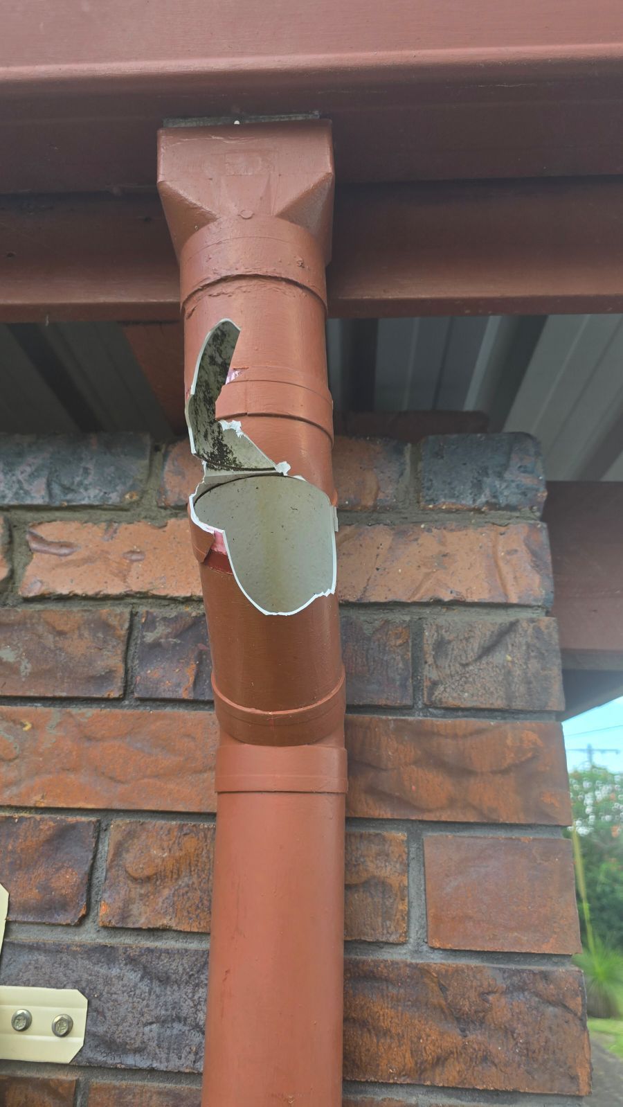 A damaged, dark-brown metal downspout with a large, jagged hole in its upper section against a brick wall.