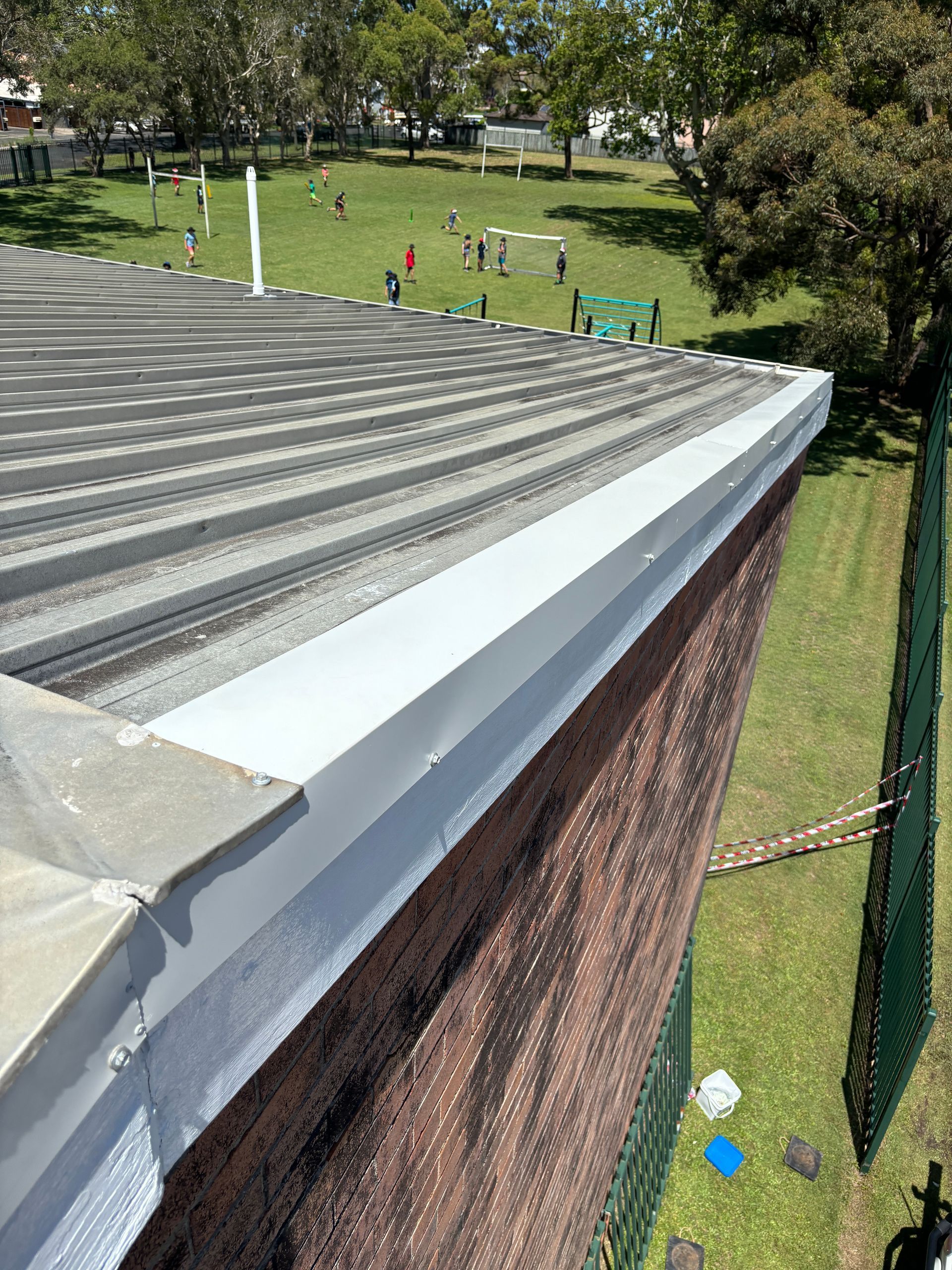An elevated view of a flat, corrugated metal roof with white flashing, looking down at a grassy schoolyard with people.