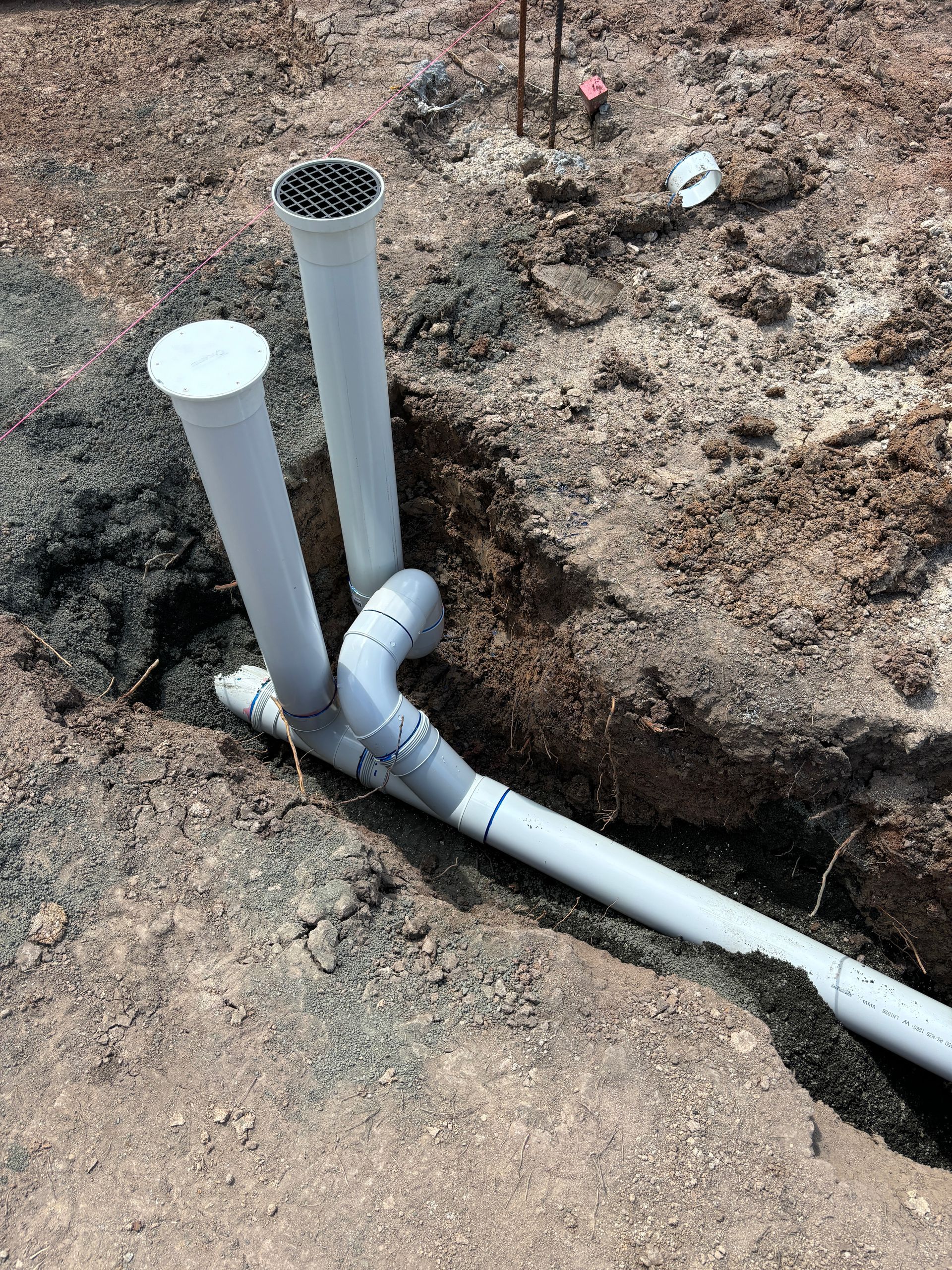 Two gray PVC pipes rise from a trench in the dirt, connected to a horizontal drainage line by an angled fitting.