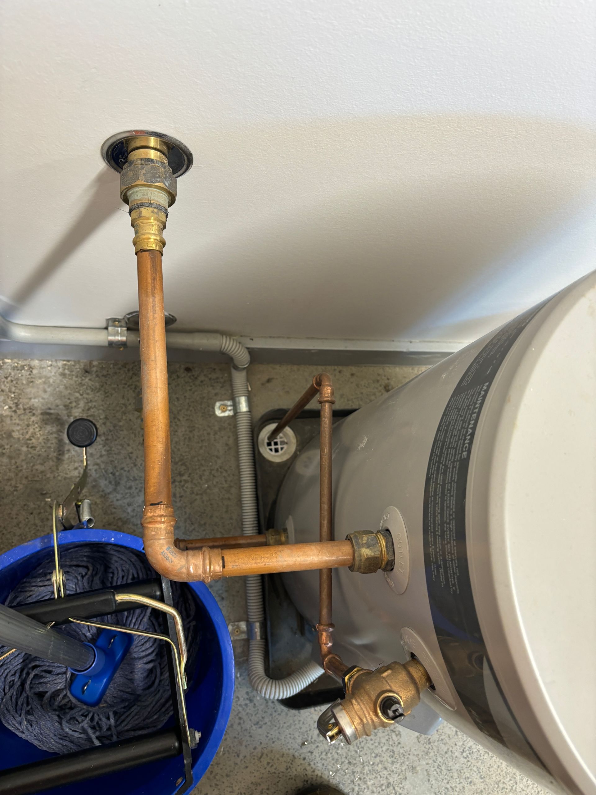 A hot water tank with copper piping connections and a pressure relief valve, viewed from above next to a blue bucket.