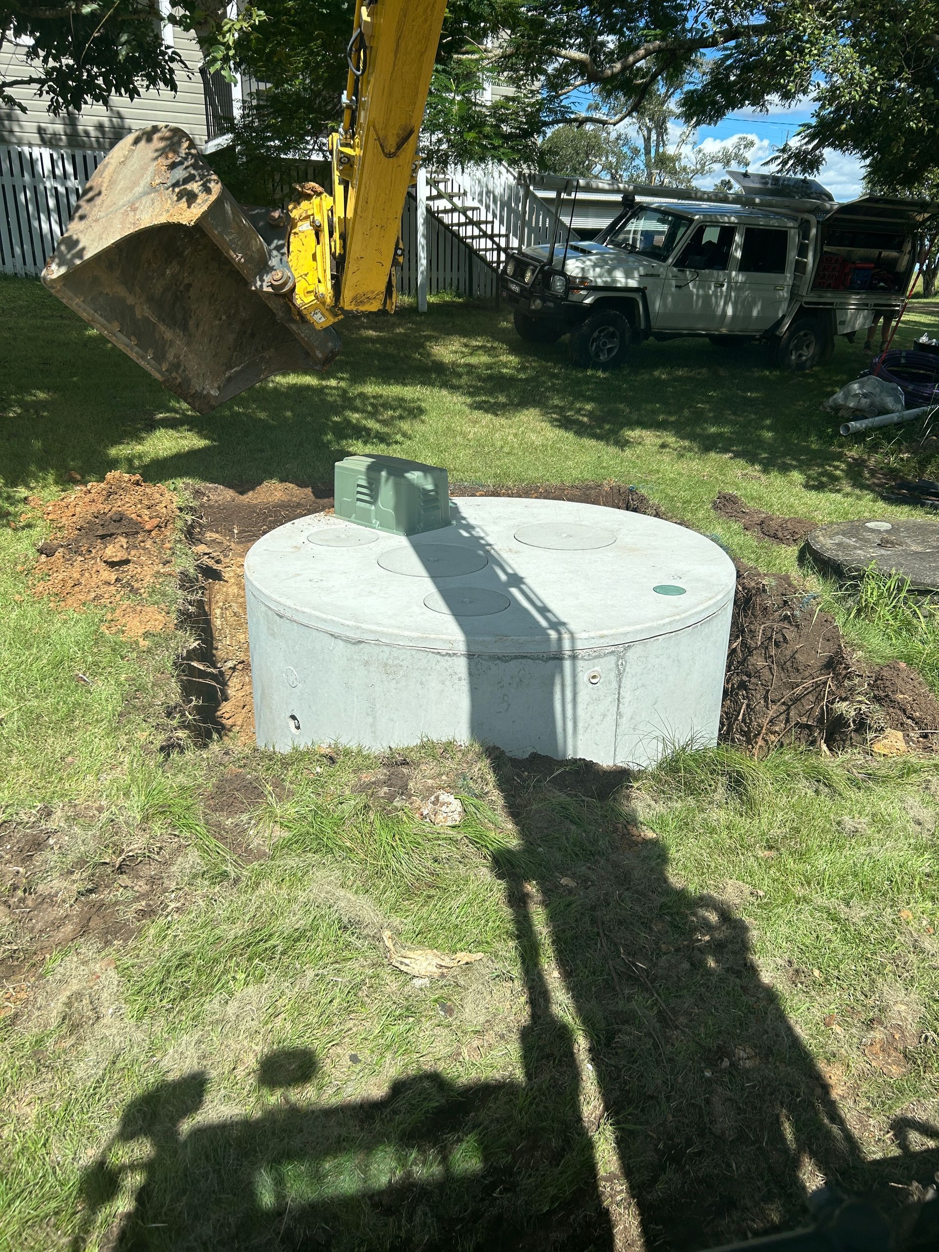 A yellow excavator arm hovers over a newly installed circular concrete septic tank in a grassy residential yard.