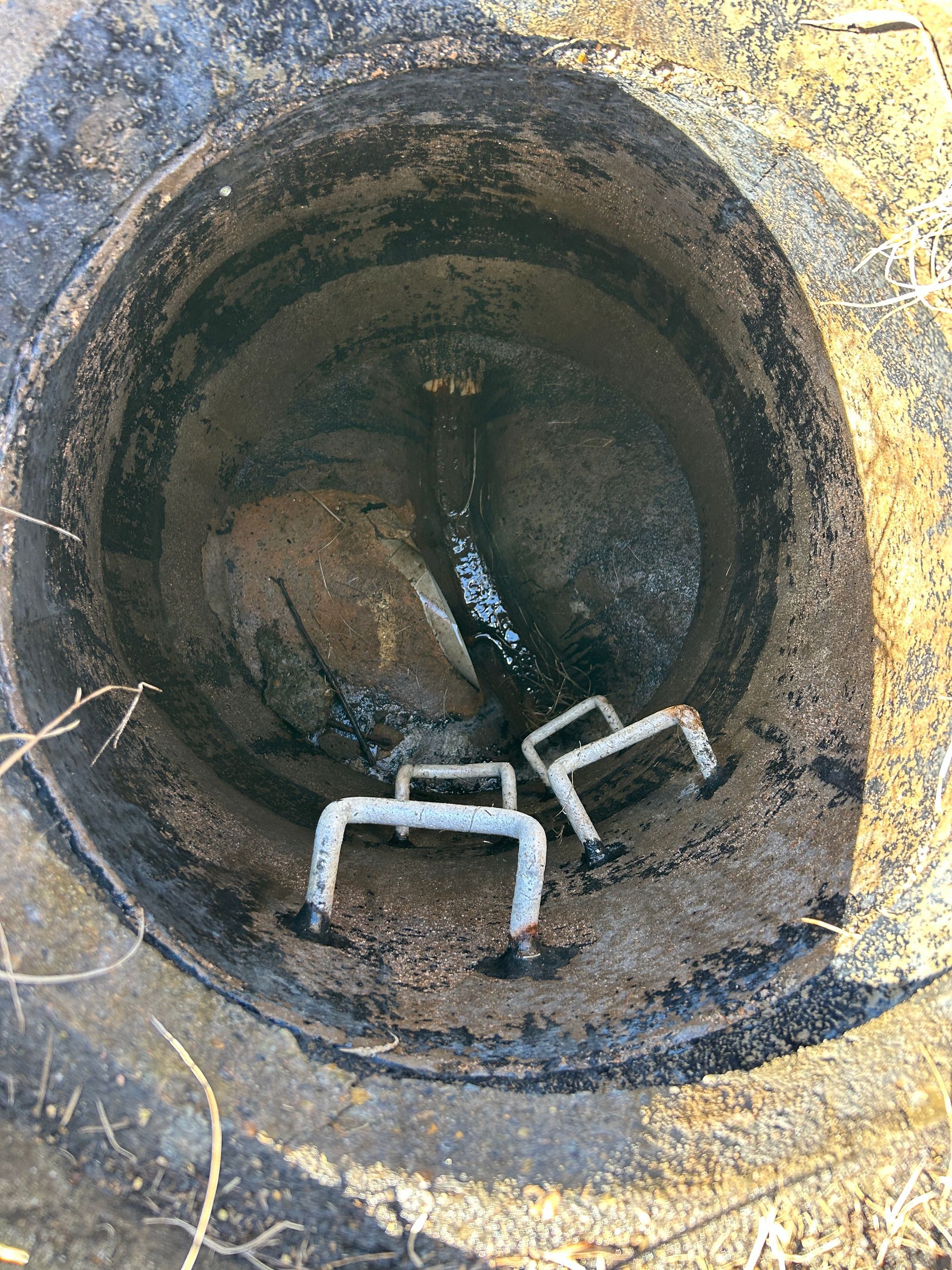 An open concrete manhole revealing a dark interior with metal steps along the wall and a pipe at the bottom.