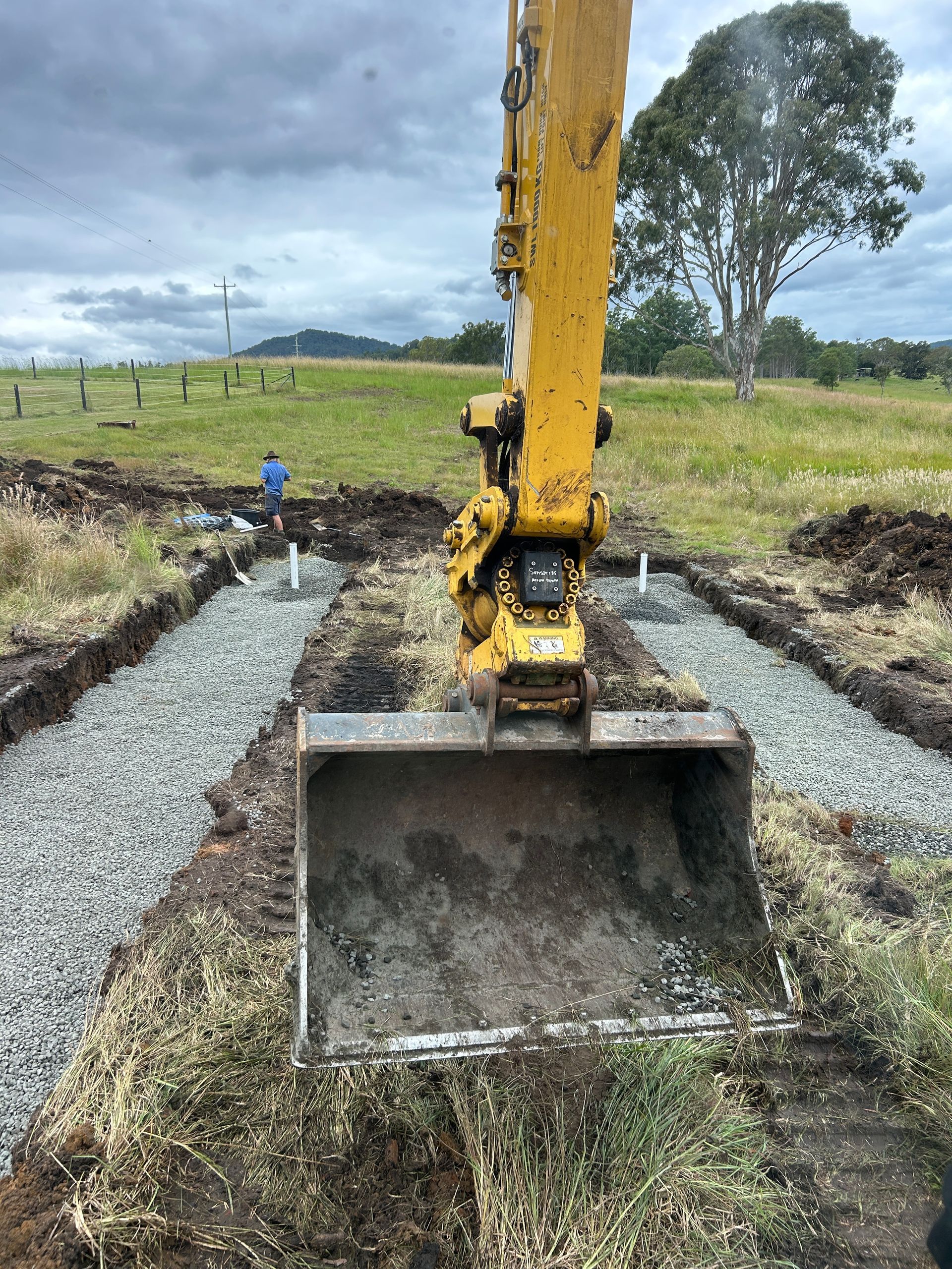 A yellow excavator bucket rests over two gravel-filled trenches in an open, grassy field under a cloudy sky.