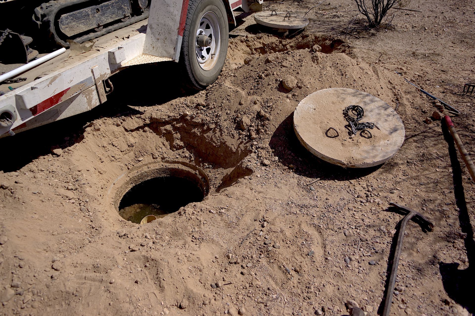 A service vehicle parked near an open septic tank lid and a hole in the sandy ground in an outdoor, arid setting.