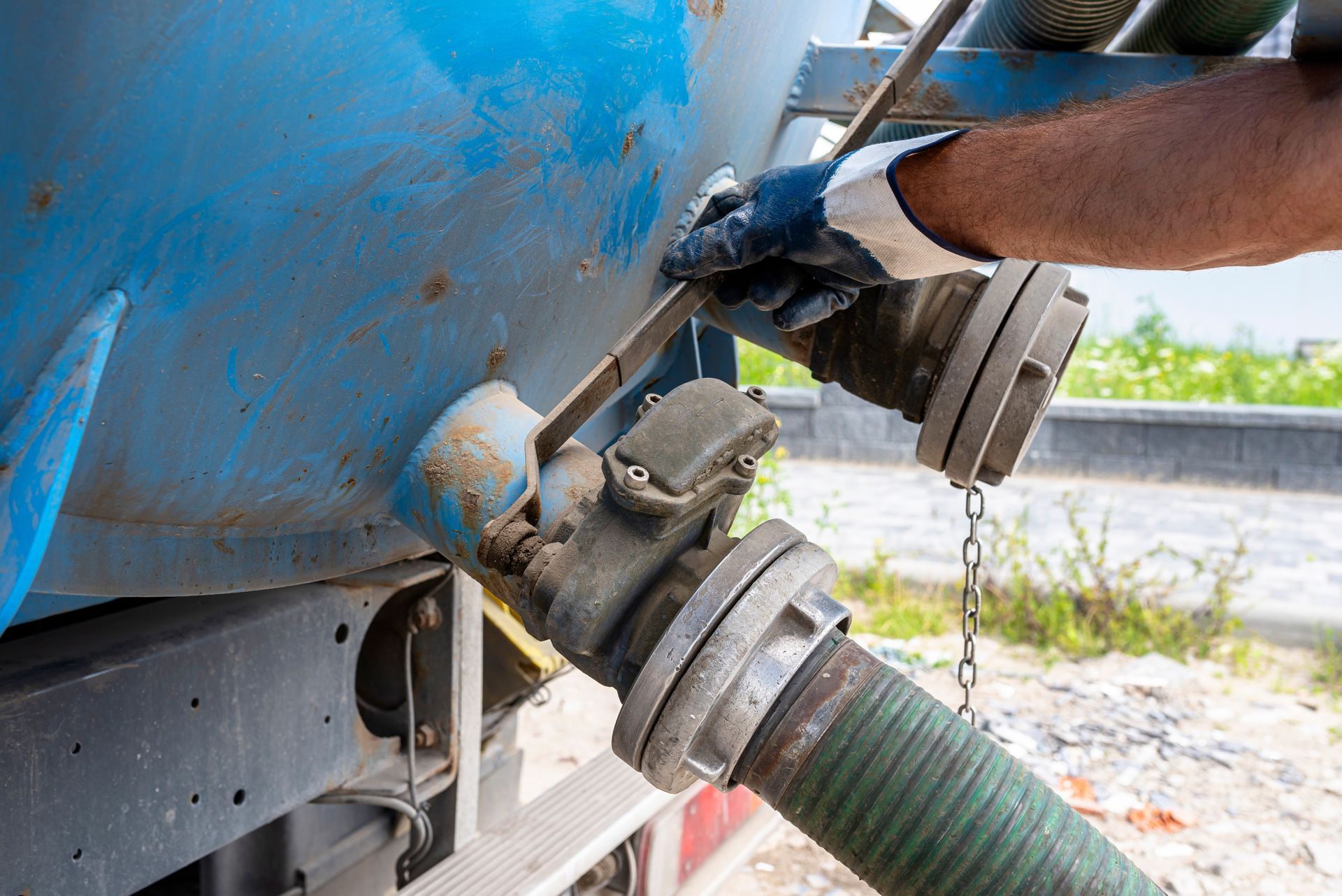 A gloved hand turns a metal valve lever on a blue industrial tank connected to a green hose.