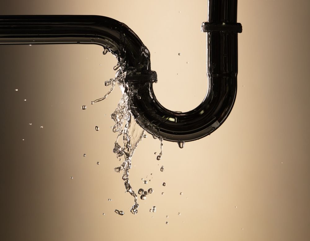 Water leaks from the joint of a dark-colored metal U-shaped pipe against a neutral background.