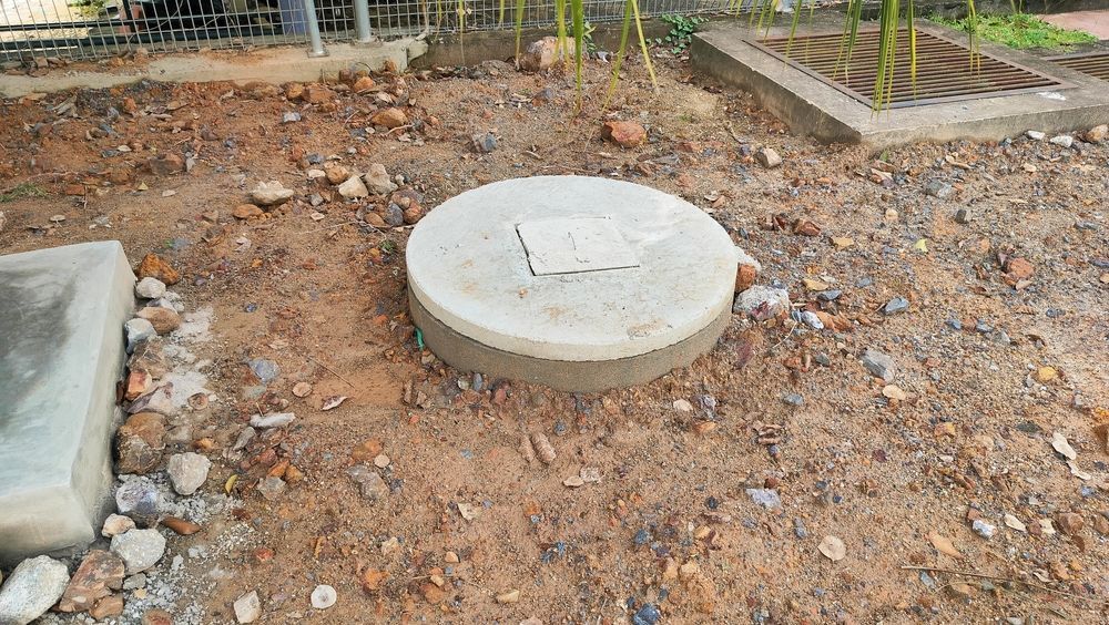 A round concrete manhole cover sits in a dirt patch surrounded by gravel and nearby storm drains.