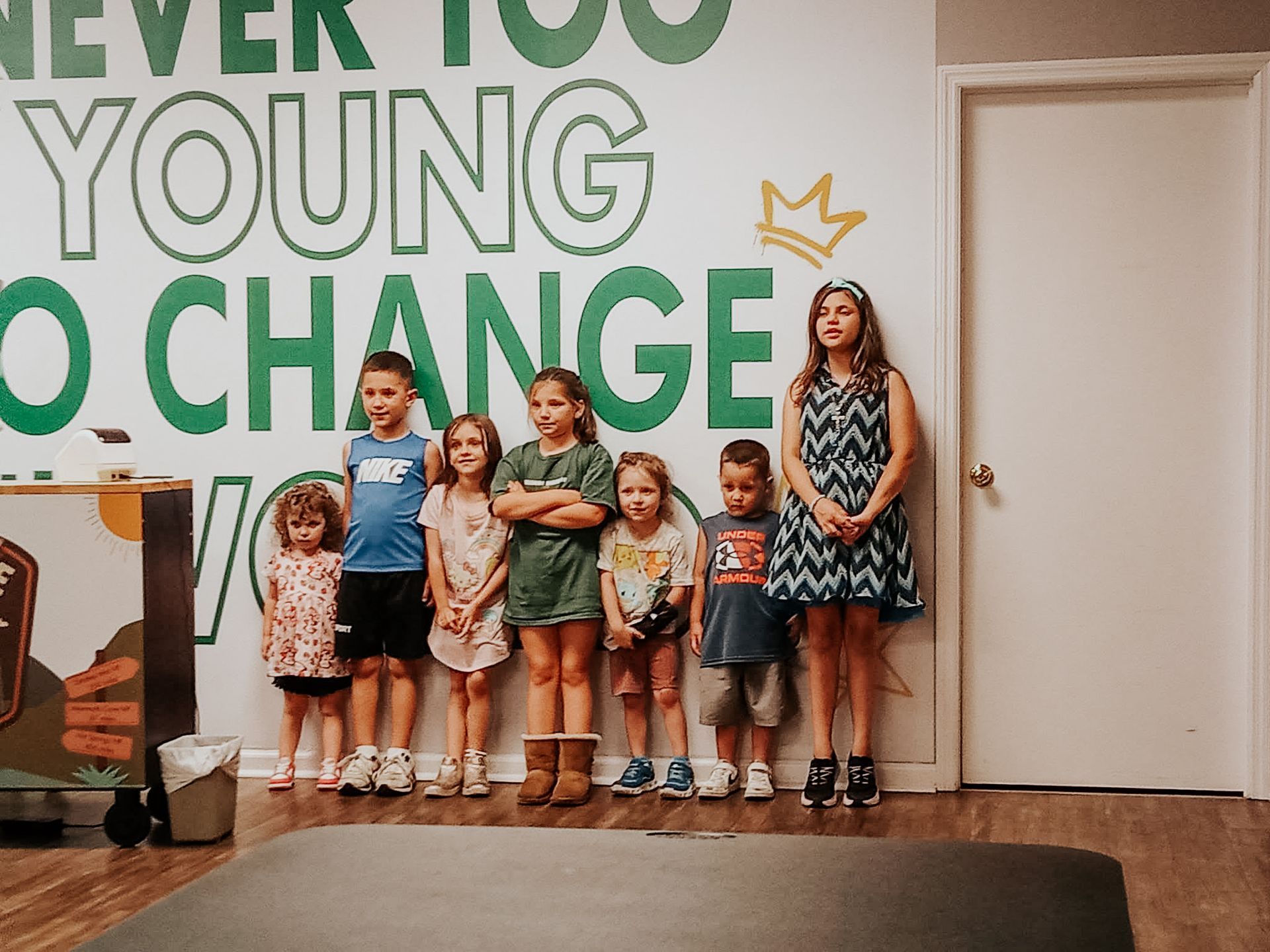 A group of children are standing in front of a wall that says `` never too young to change ''.