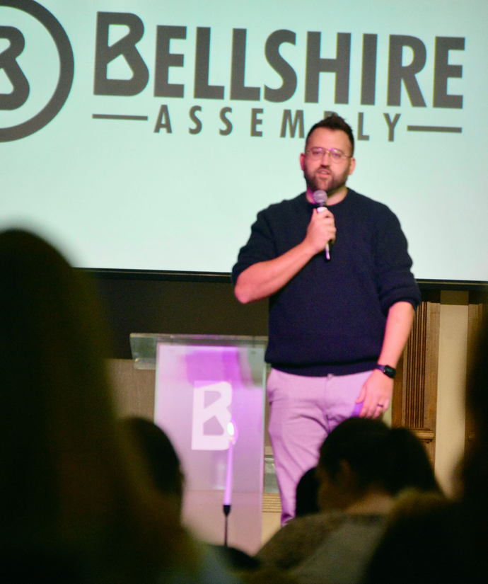 A person speaks into a microphone at a podium in front of a projection screen that reads, 