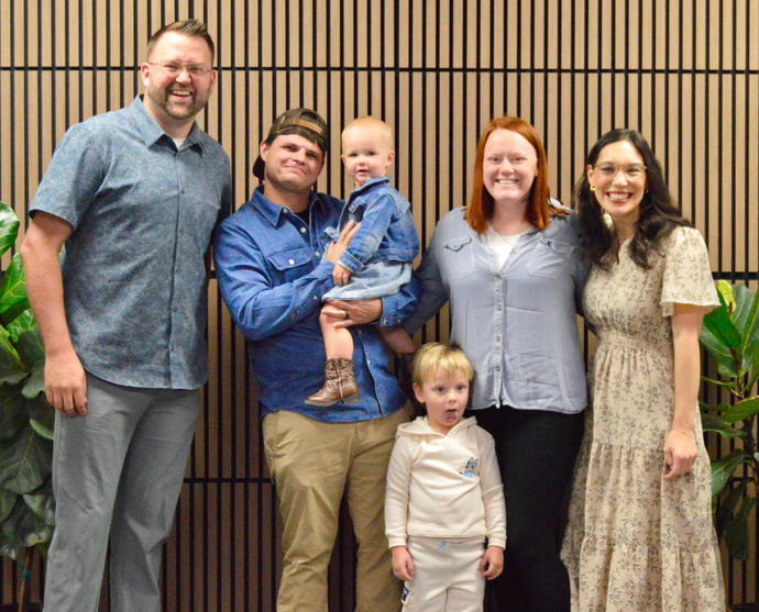 Group portrait of six people standing in front of a wooden slat wall with indoor plants on either side.