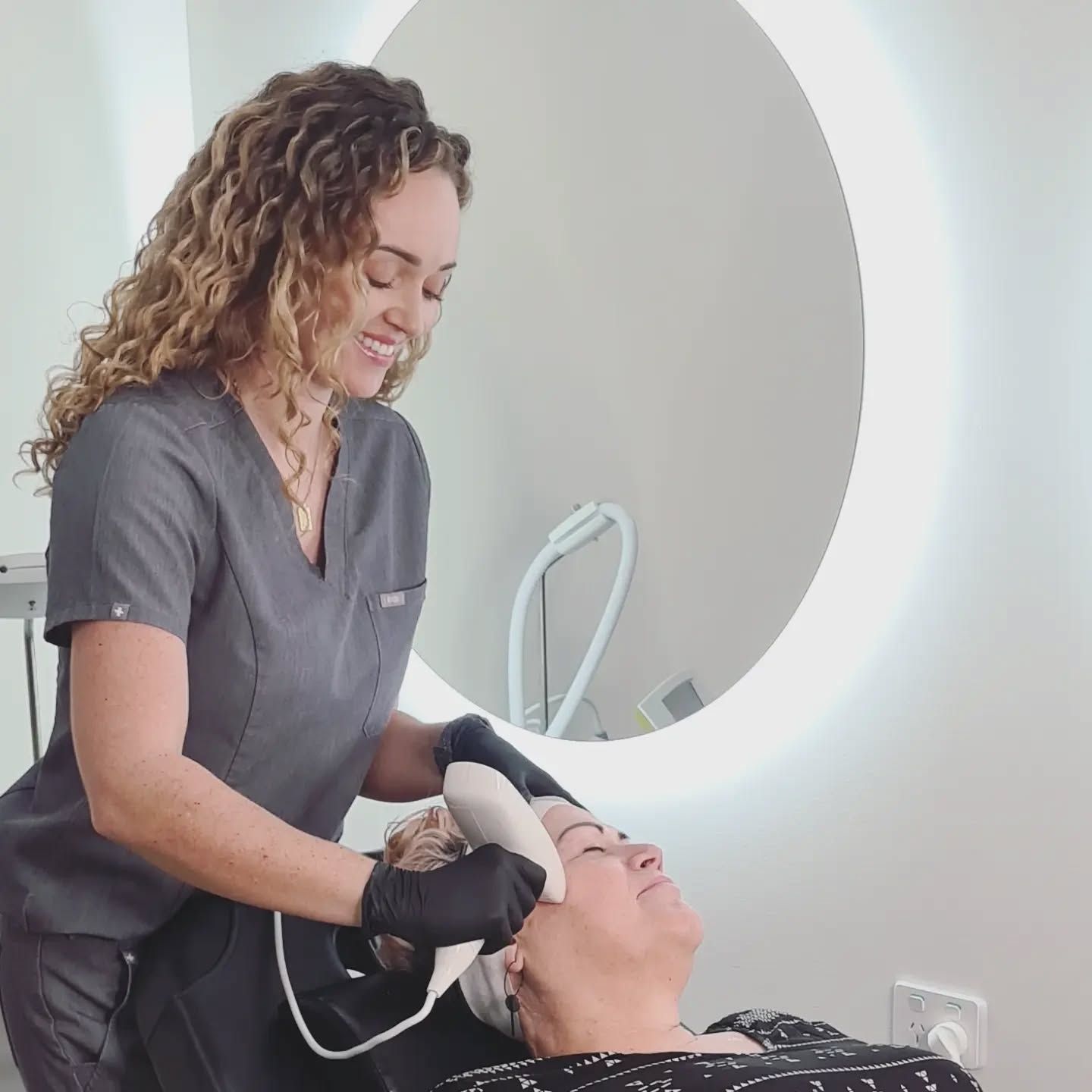 A Woman is Giving a Woman a Facial Treatment in Front of a Mirror — Nurose Aesthetics in Nundah, QLD
