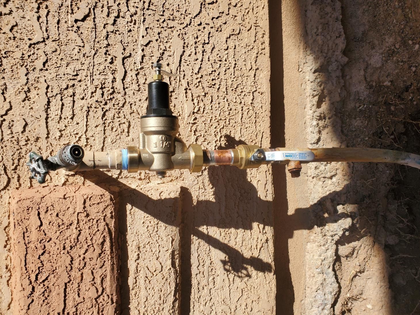 A water pressure regulator and connected pipes mounted on a stucco wall outside.