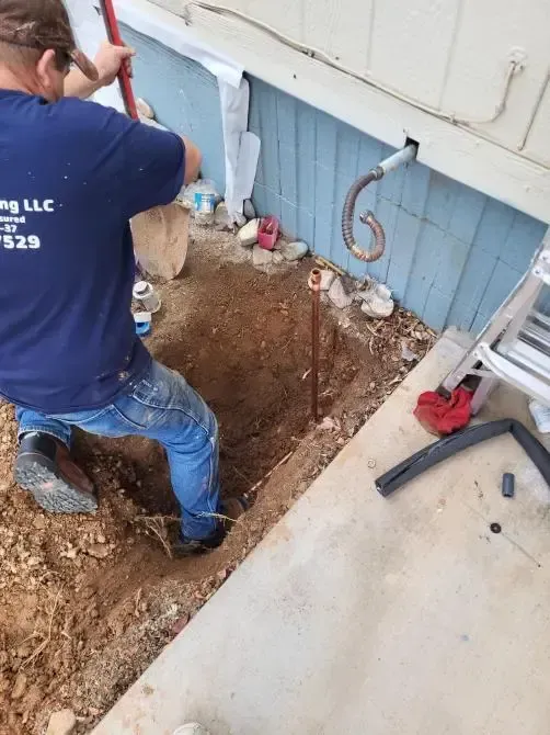 Person digging near a building's foundation. Exposed pipe, dirt, and tools present. Blue siding.