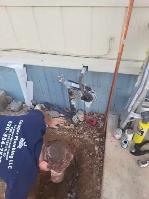 Plumber repairing pipes near a building's exterior. Copper pipe, black drain pipes, and dirt visible.