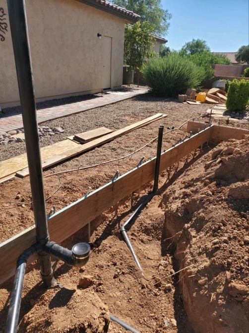 Construction site: wooden forms, plumbing, and dirt. A trench is visible with pipes, along with a house in the background.