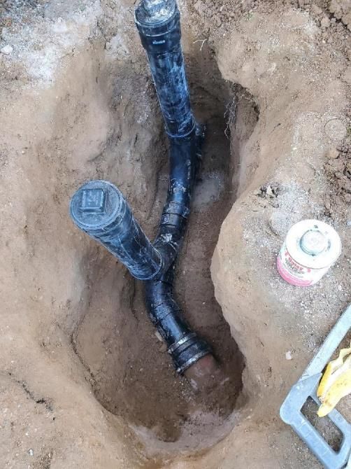 Black plumbing pipes in a trench, likely being installed. There is a can of glue and a level tool nearby.
