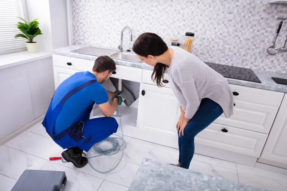 Plumber fixing kitchen sink as a woman watches. White cabinets, tiled backsplash, tools, blue uniform.