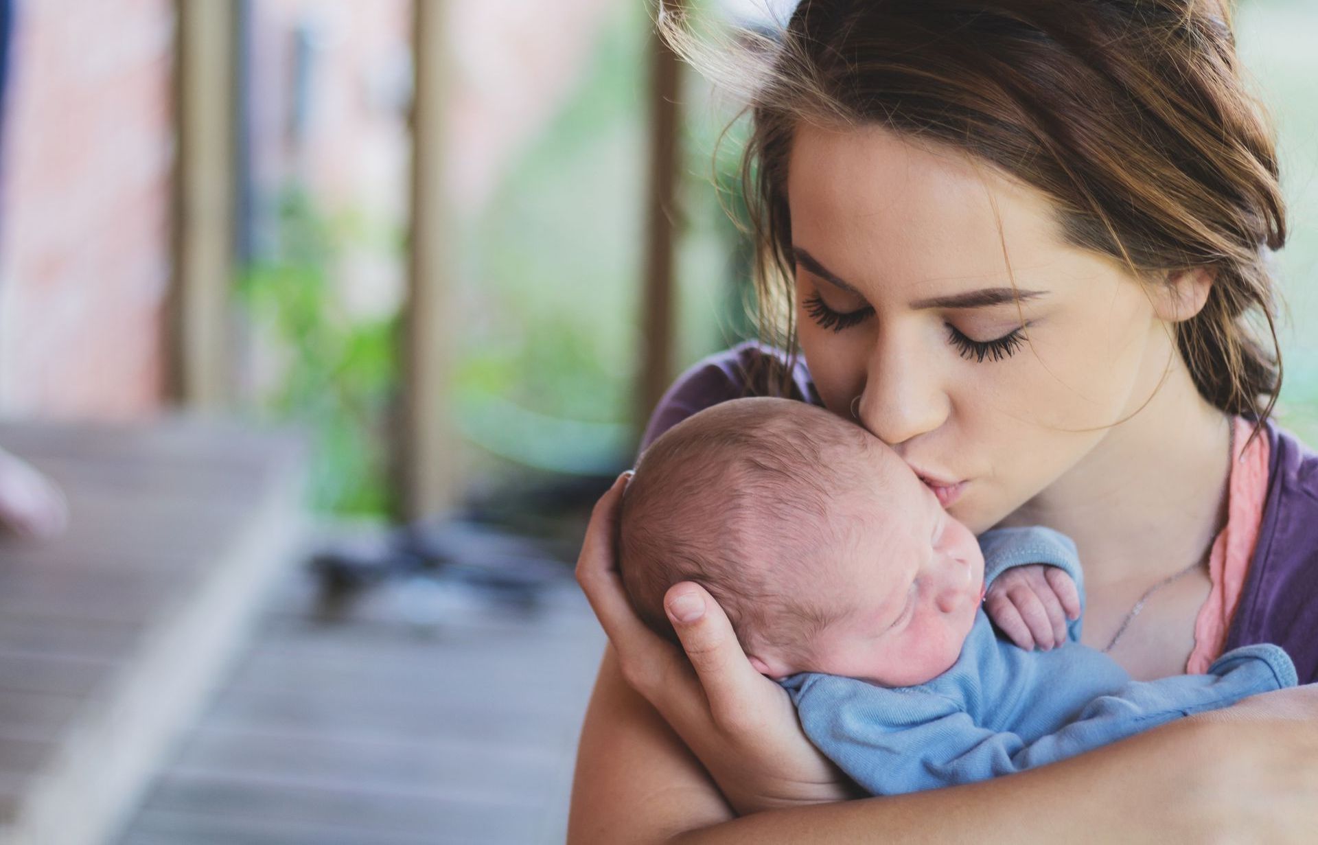 Woman kisses baby's head outdoors; baby in blue onesie, woman in purple top.