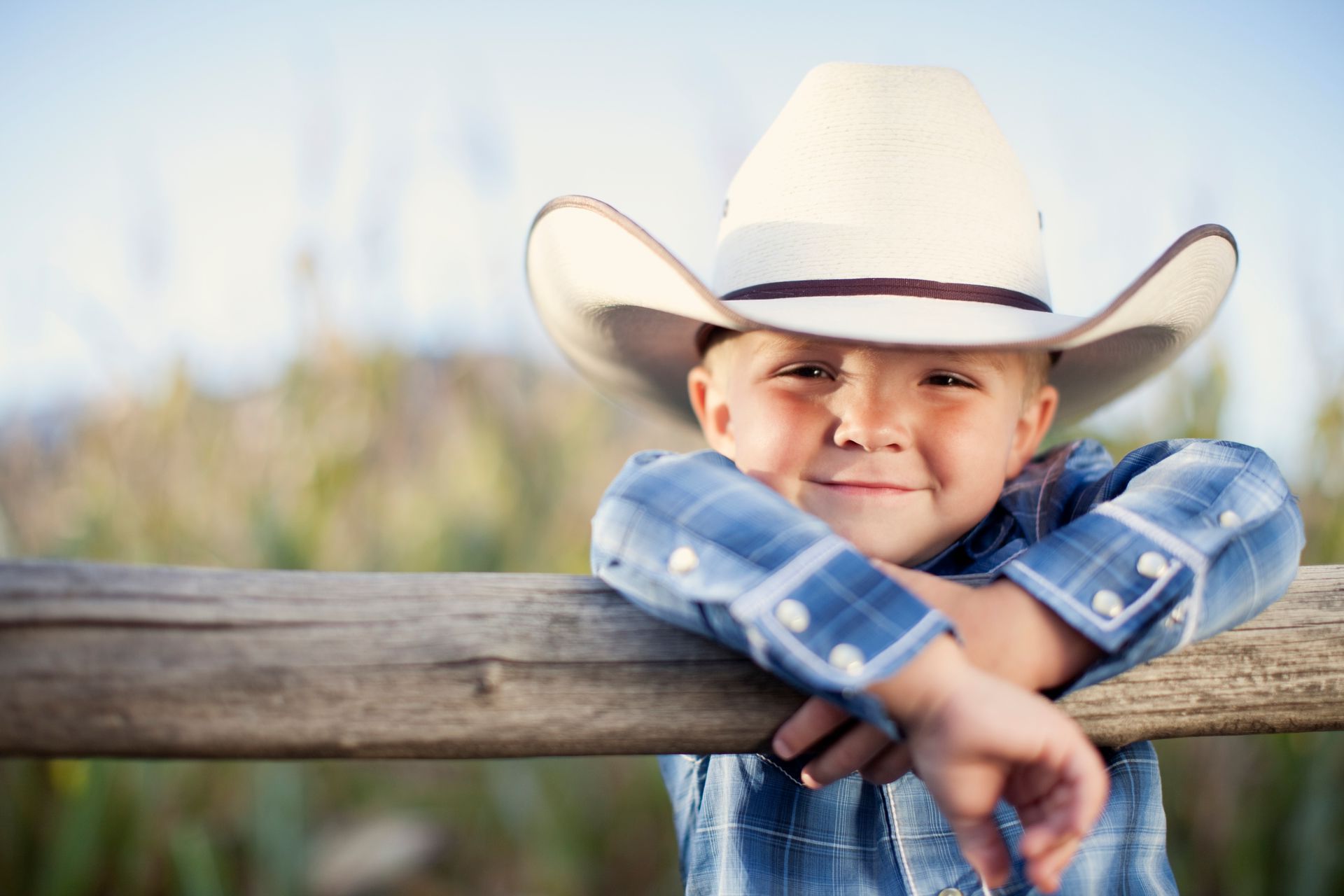 A young boy wearing a cowboy hat leans on a wooden fence.