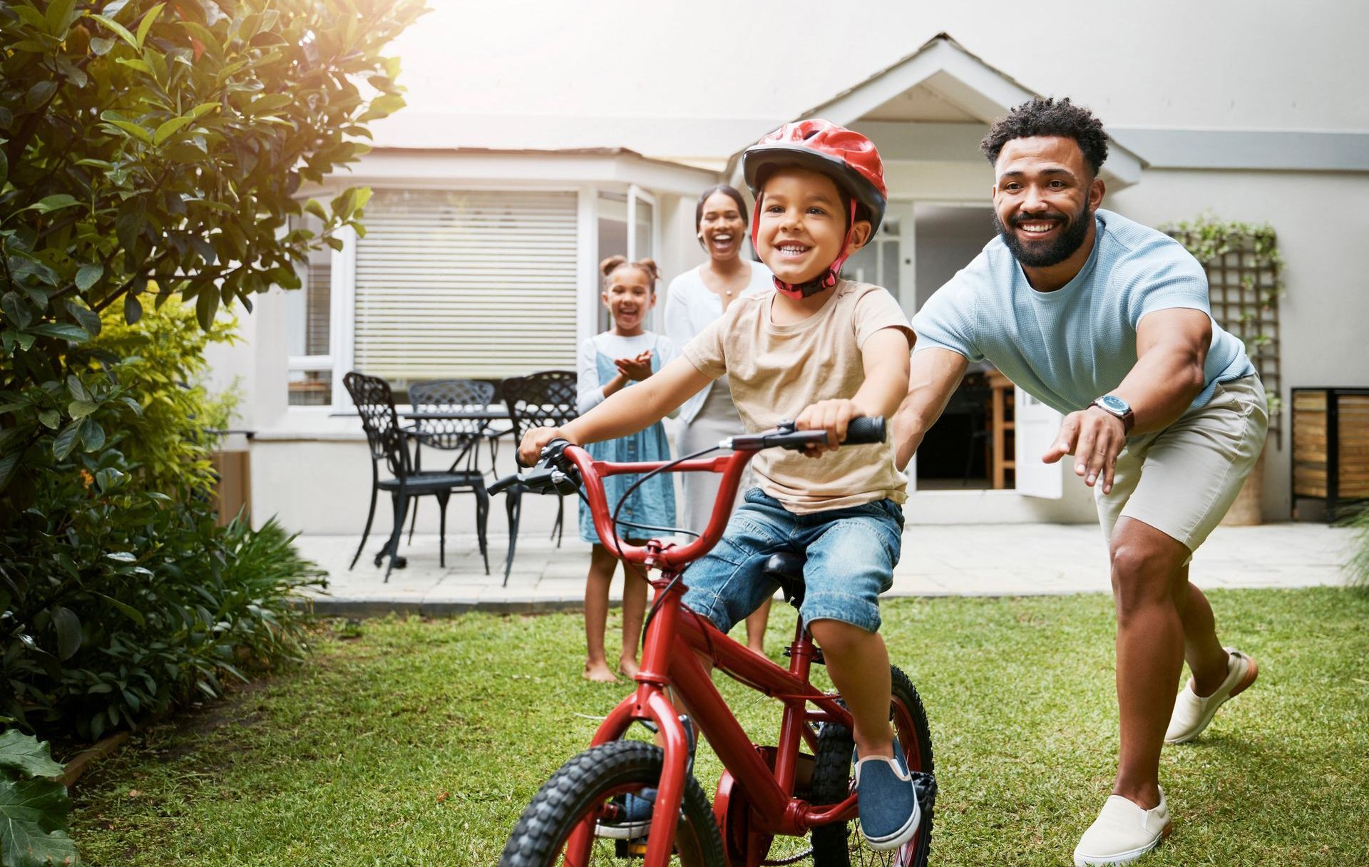 A person helping a child on a red bicycle in a backyard, with others watching in the background.