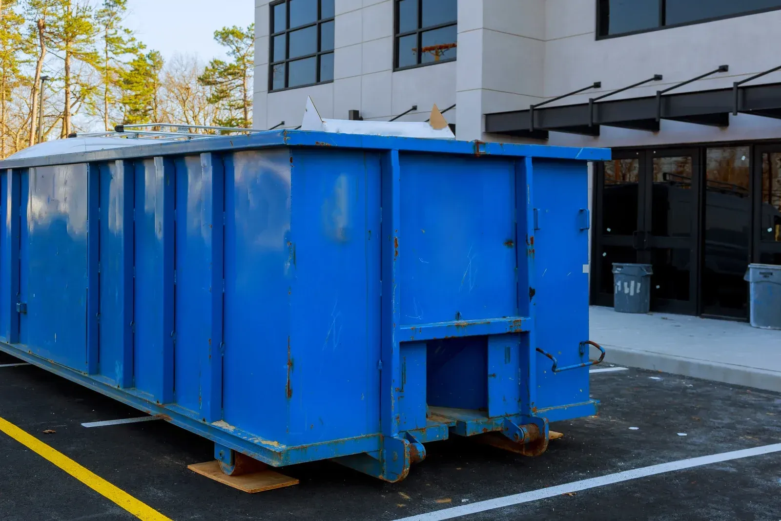 Blue dumpster in front of a modern building with black doors and windows, parked on asphalt with yellow lines.
